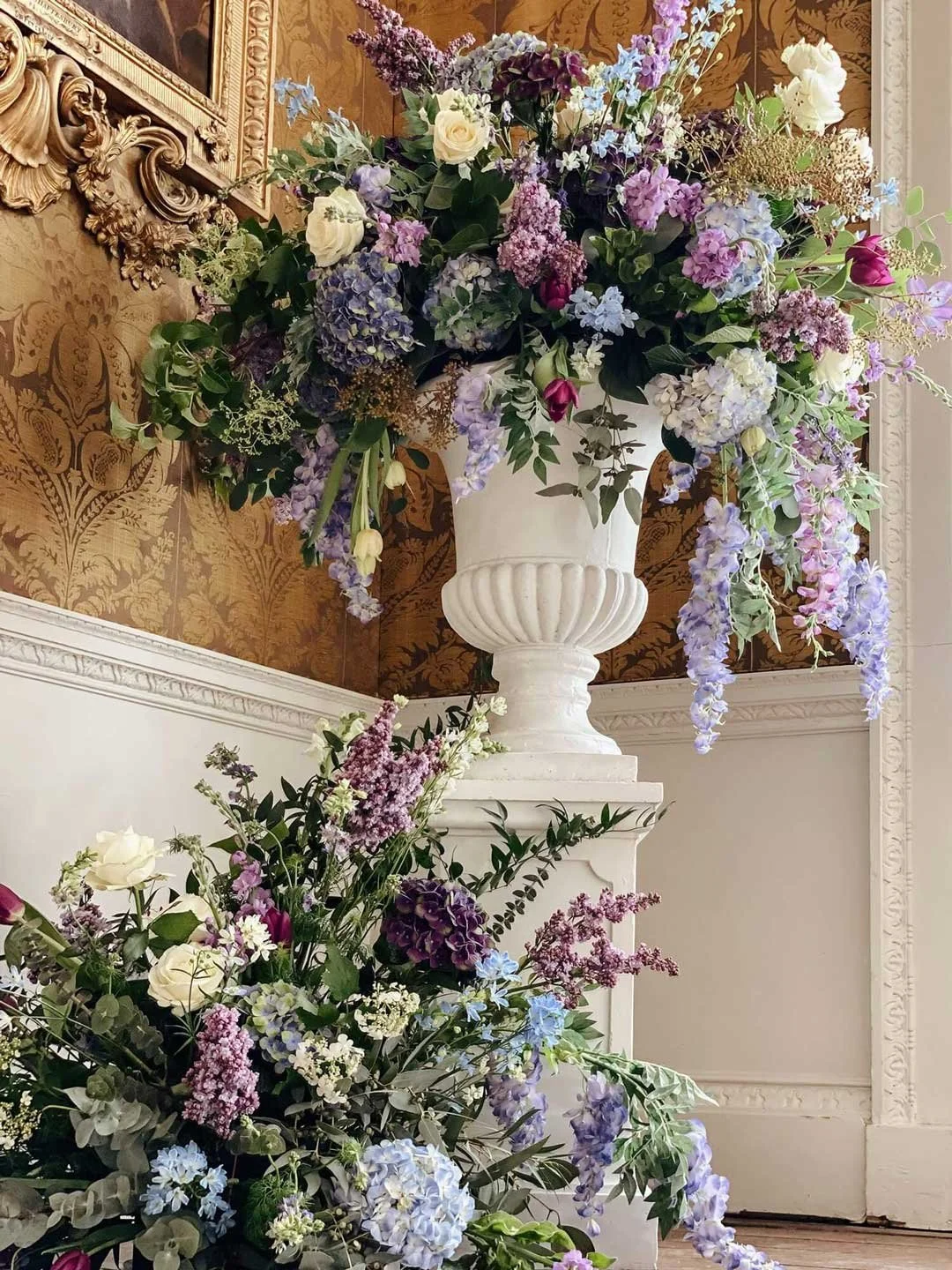 Large floral arrangement with purple, white, and pink flowers in a white urn-shaped vase, placed atop a pedestal in a room with ornate gold and white decor.
