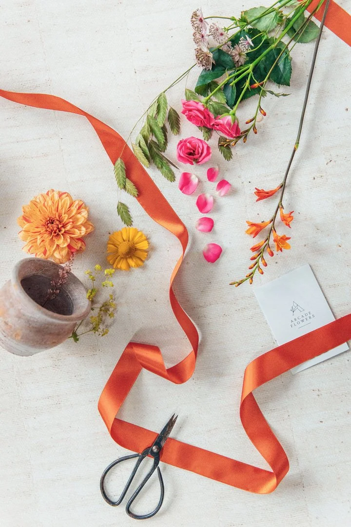 Arrangement of pink, yellow, and orange flowers with green foliage, orange ribbon, scissors, a small pink card with logo, and a ceramic vase on a white wooden surface.