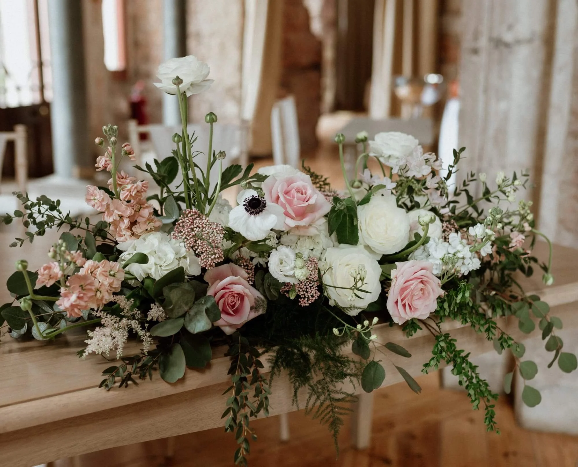 A floral arrangement with pink and white roses, anemones, and greenery on a wooden table in a rustic indoor setting.