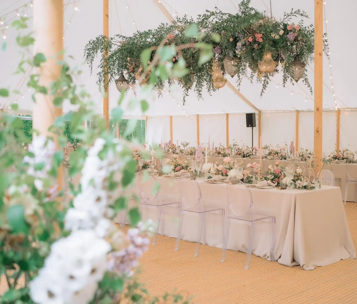 A wedding reception in a marque is decorated with beautiful wedding flowers. Flowers are seen on the tables and hanging from the ceiling.
