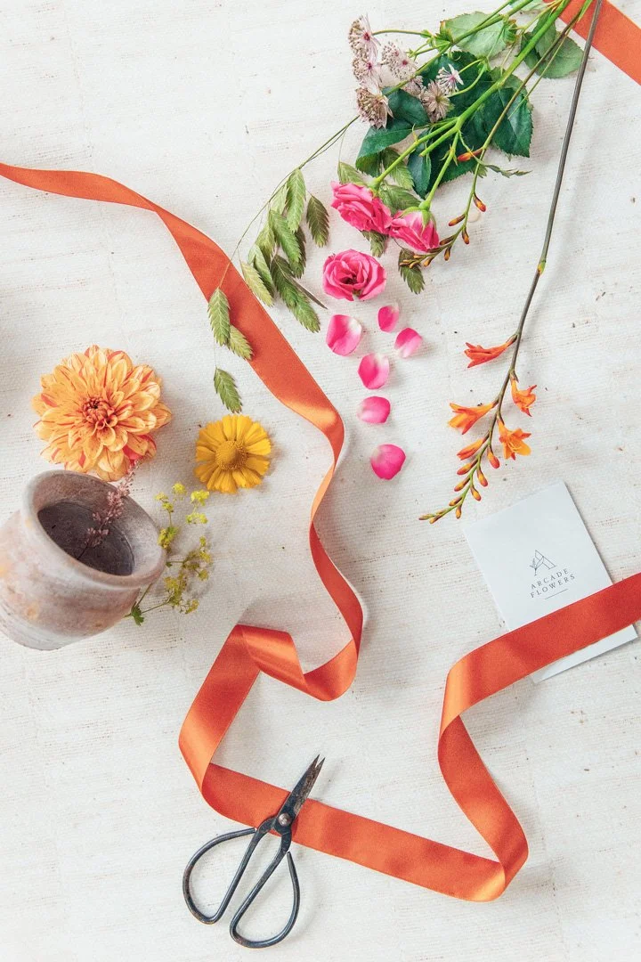An overhead view of a florist’s workshop table showing fresh flowers, a ribbon, some scissors and a gift tag.
