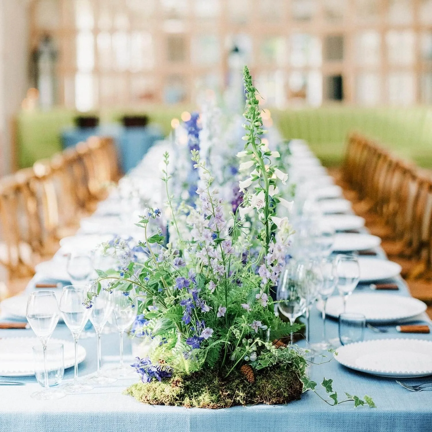 Table decorated with a tall flower arrangement of purple, blue, and white flowers on a light blue tablecloth, set for a formal event with plates, wine glasses, and silverware in a bright room with large windows.