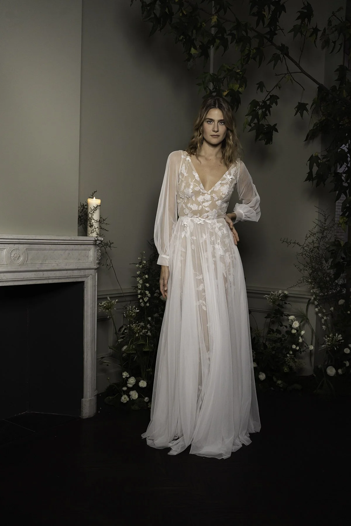 Woman wearing a lace wedding dress with tulle skirt in a dark room with candle, trees and a fireplace
