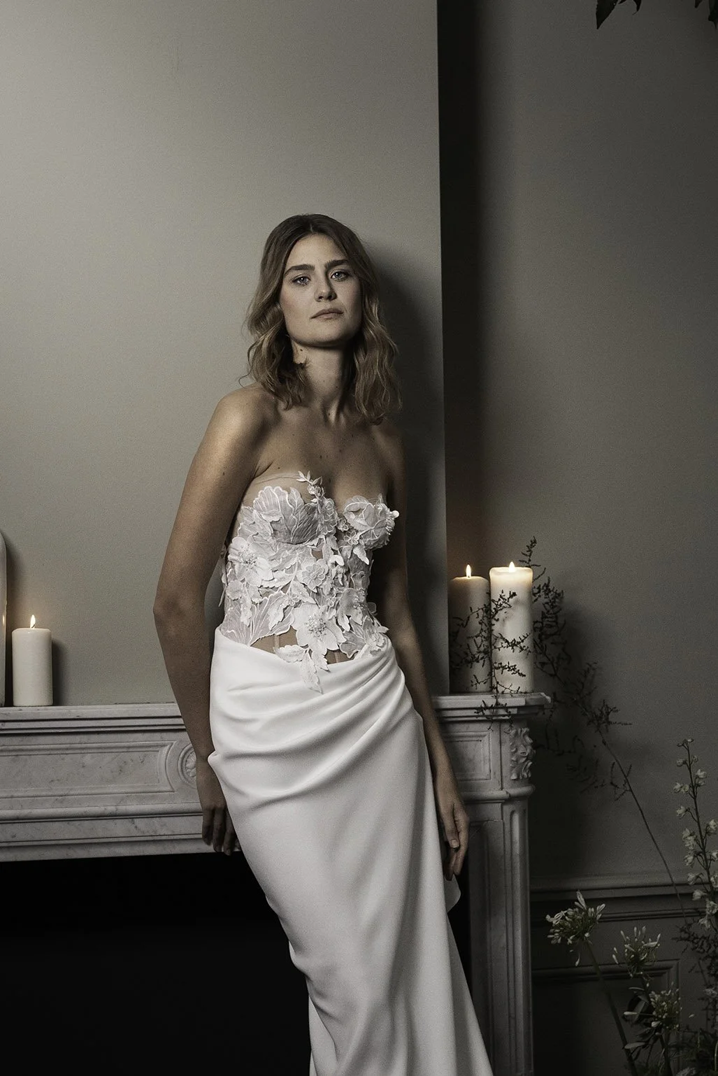 Woman wears wedding gown with 3D lace corset and draped crepe skirt leaning against a fireplace