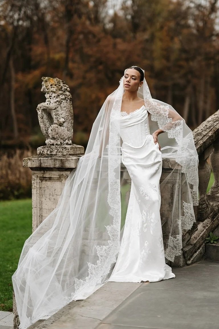 Bride in a striking Edwin Oudshoorn white wedding dress made from floral jacquard satin fabric and veil standing outdoors next to stone sculptures and stairs, with a background of trees with autumn leaves.