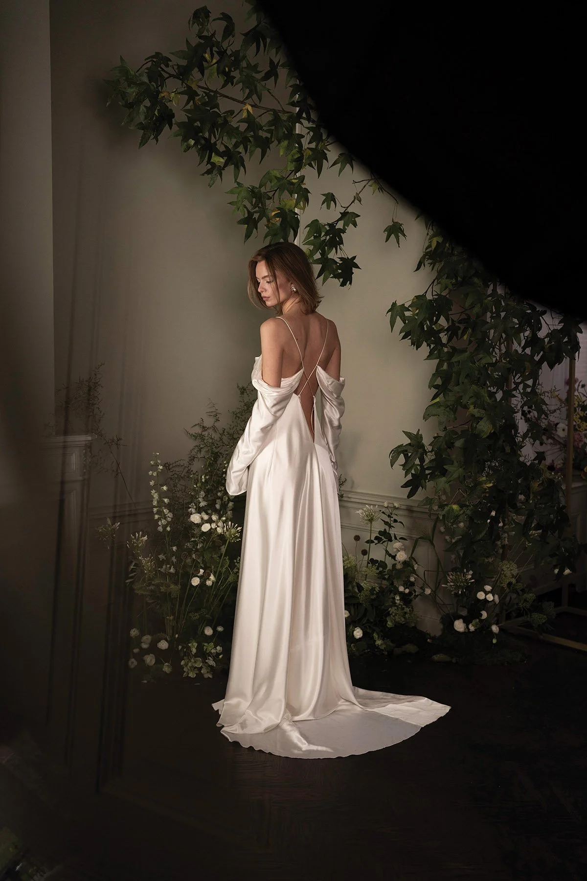 Woman wearing a draped satin silk bias cut dress with deep open back and long sleeves in a room witha. tree