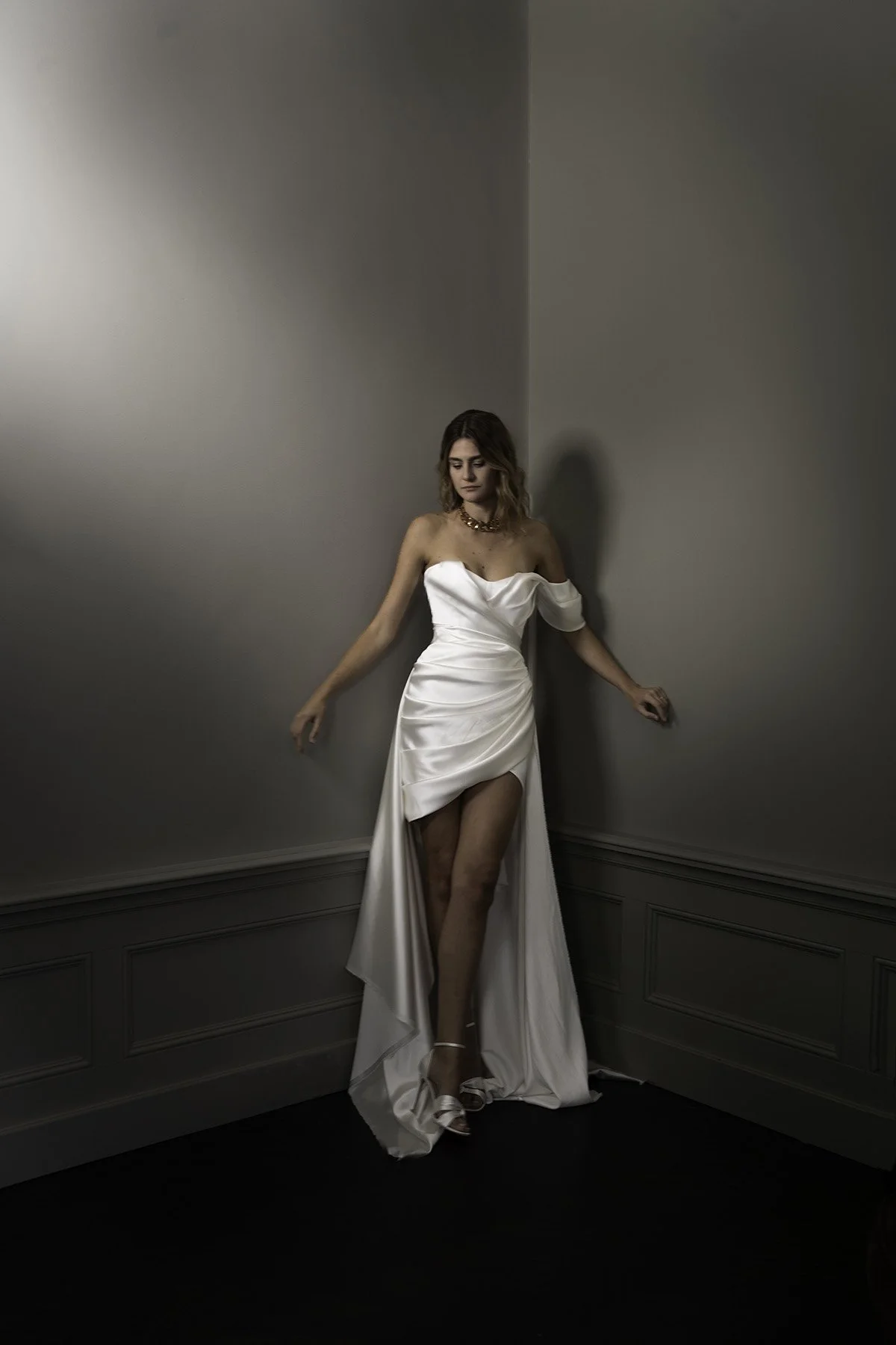 Woman stands in ivory satin short strapless dress with draped body and skirt. She wears a beaded necklace and is standing in a dark beige room