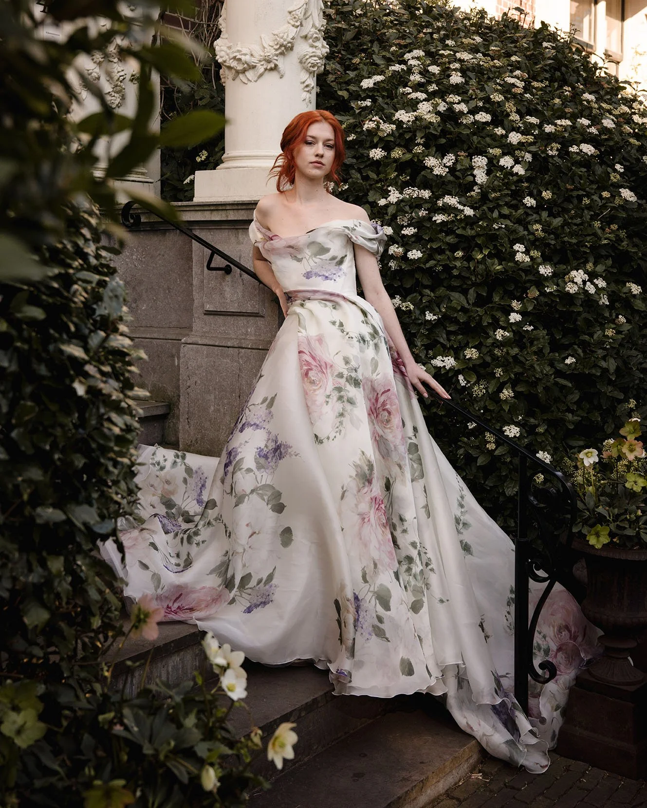 A bride with red hair wearing an Edwin Oudshoorn floral off-the-shoulder silk organza ballgown standing on outdoor stairs surrounded by greenery and white flowers.