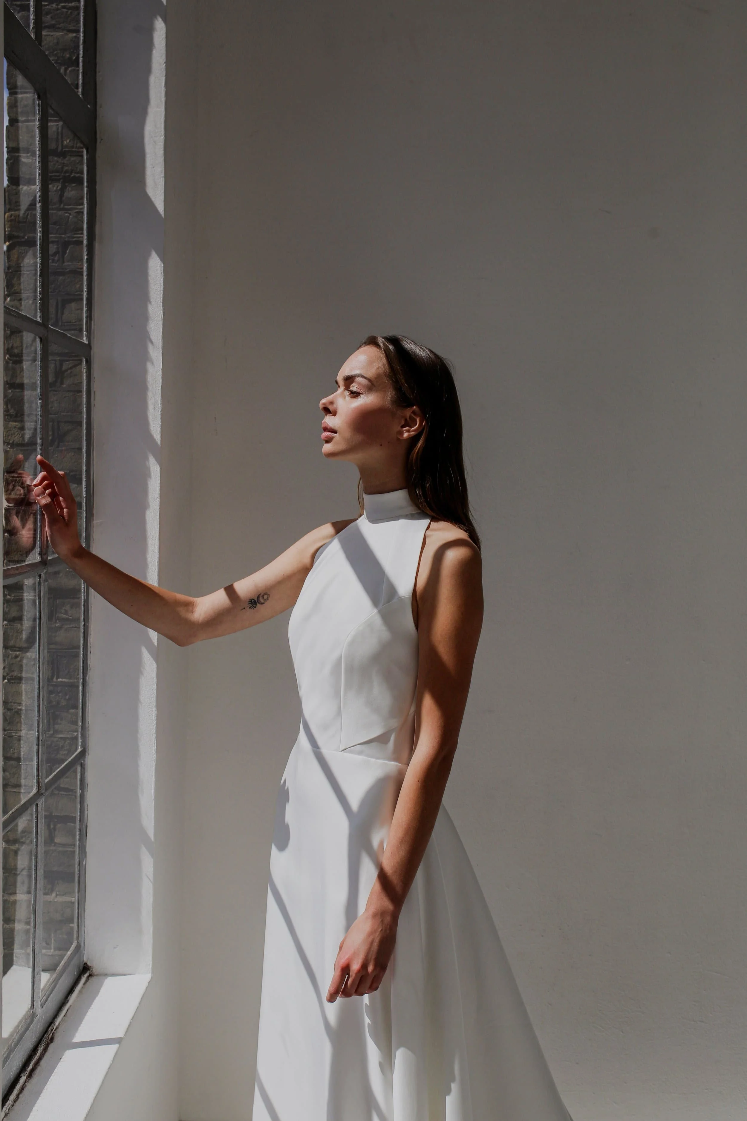 A woman in a satin halterneck wedding dress stands in a bright, minimalist white room with tall windows.