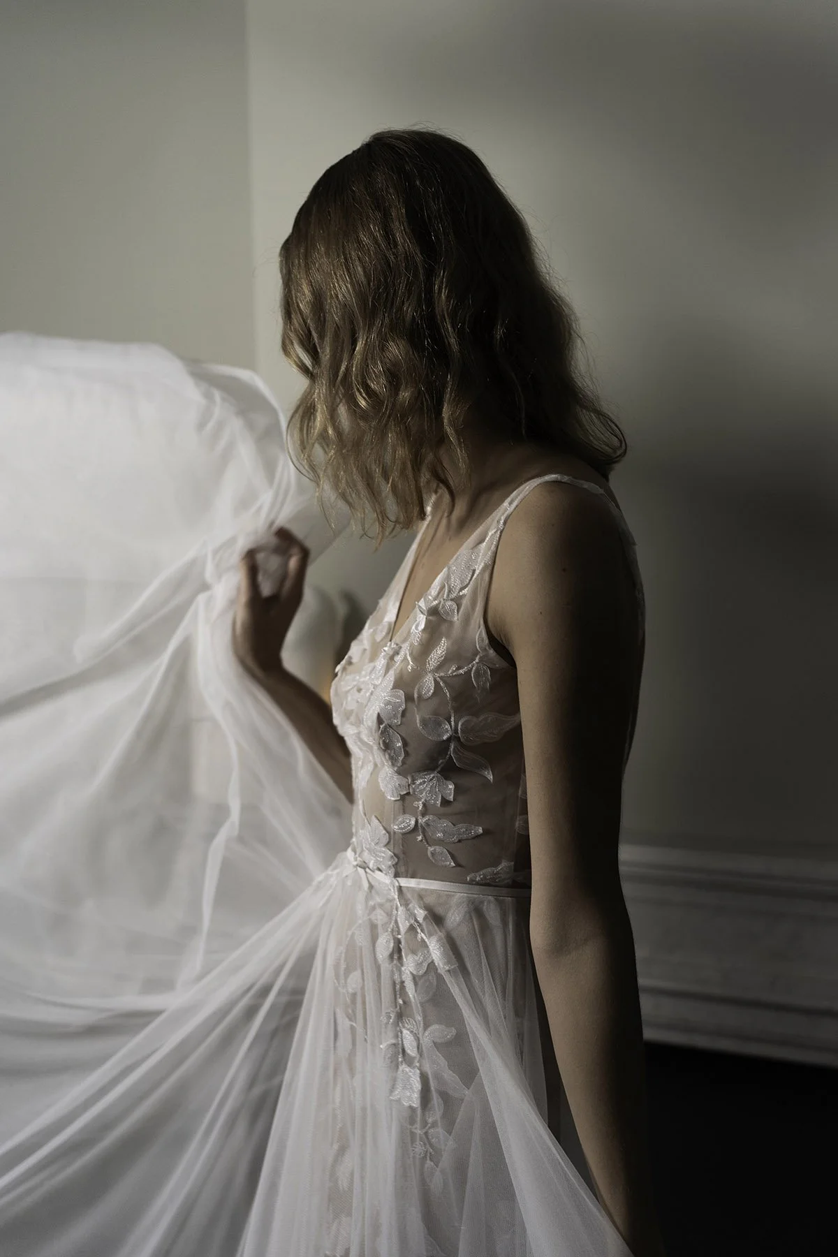 Woman wearing a lace wedding dress with tulle skirt in a dark room with candle, trees and a fireplace