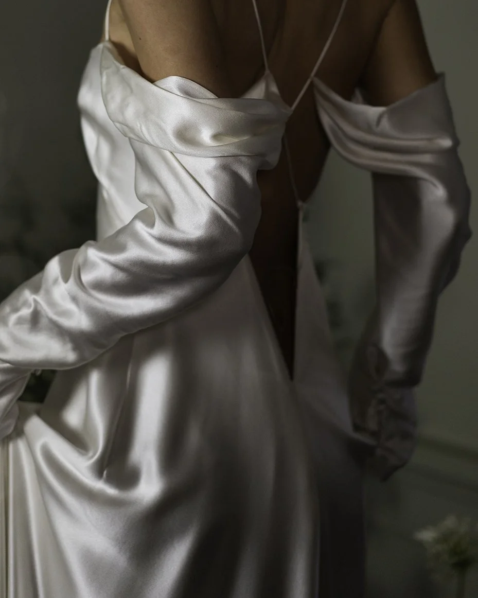 Woman wearing a draped satin silk bias cut dress with deep open back and long sleeves in a room witha. tree