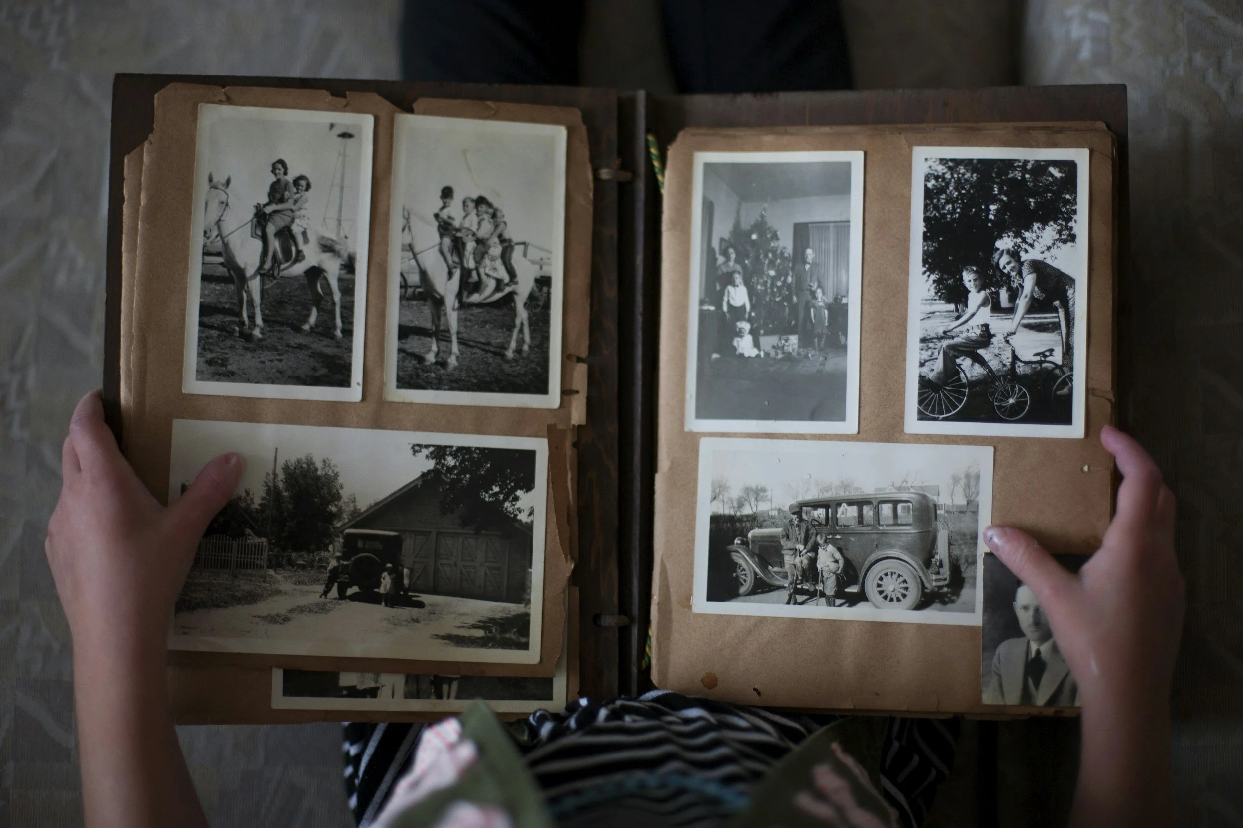 A person is holding an old photo album open to black-and-white photographs, some depicting children playing and others showing houses, cars, and people indoors.