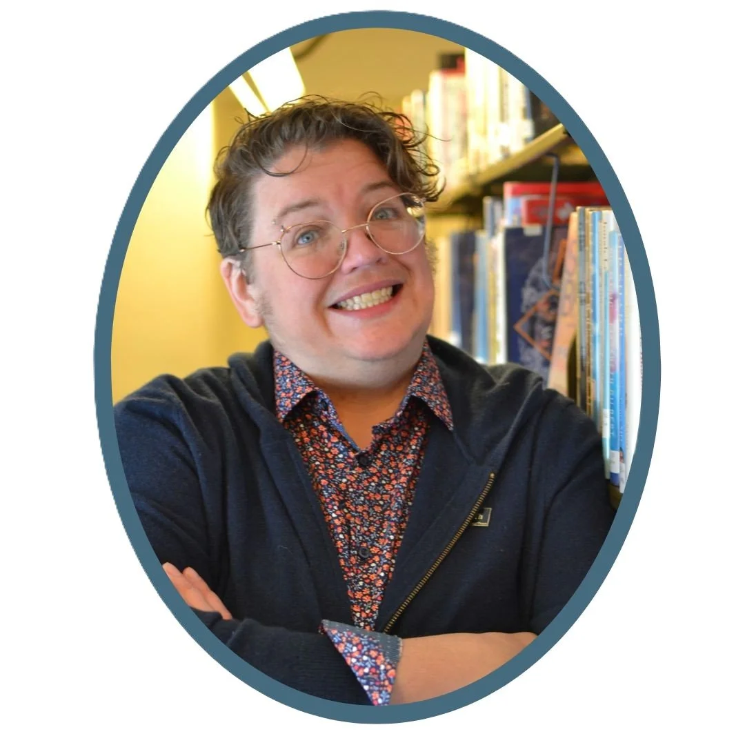 Mx. Katharine, a chubby white person in their 40s with curly brown hair and wireframe glasses, is leaning against a library bookshelf with their arms crossed.  They are grinning into the camera with a somewhat unhinged look on their face.