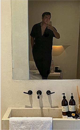 Person taking a mirror selfie in a bathroom, wearing a black outfit, with bathroom supplies and a towel visible in the foreground.