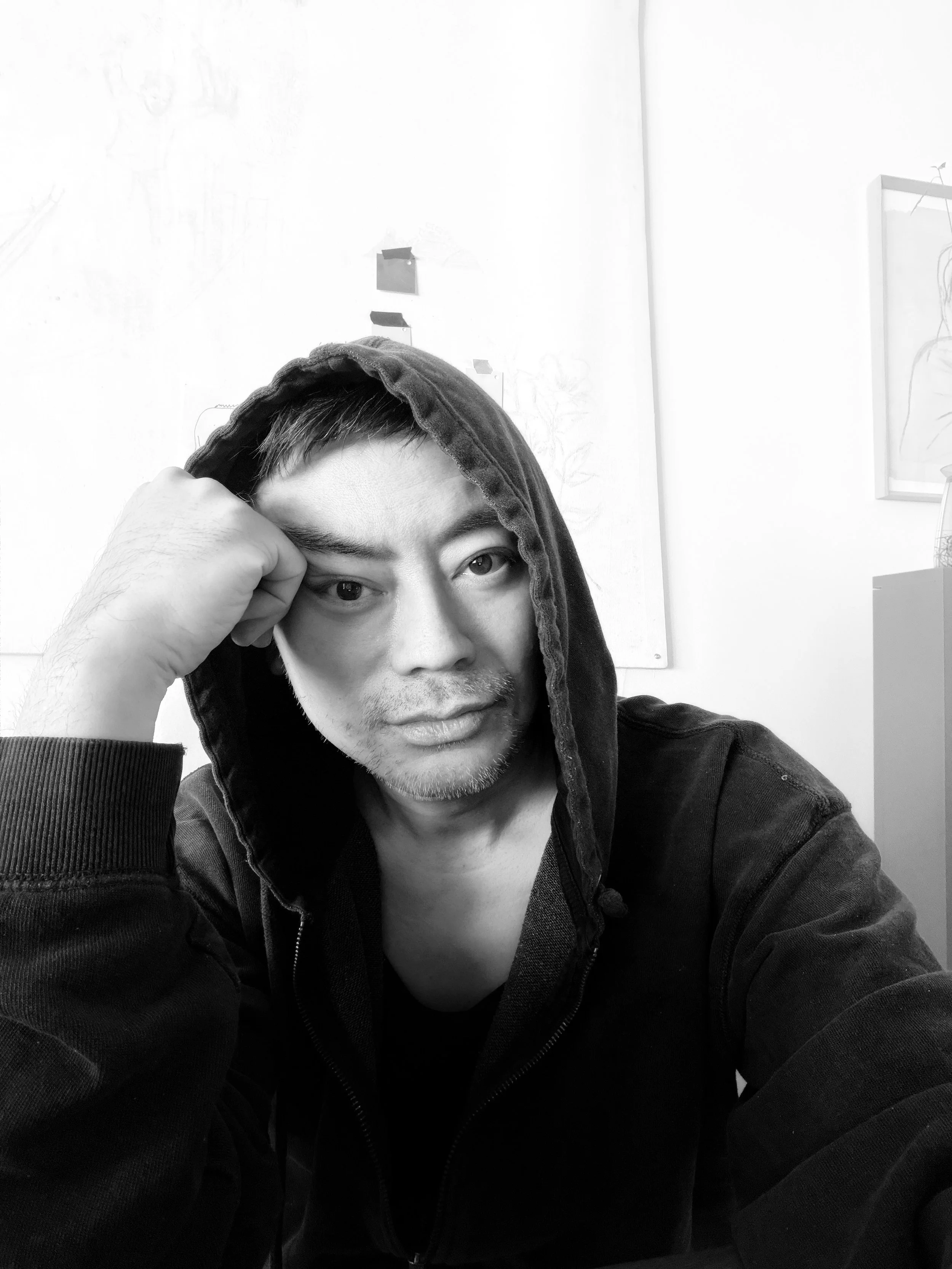 Black and white photo of a young man with short hair wearing a hoodie, sitting indoors, resting his head on his hand with a thoughtful expression.