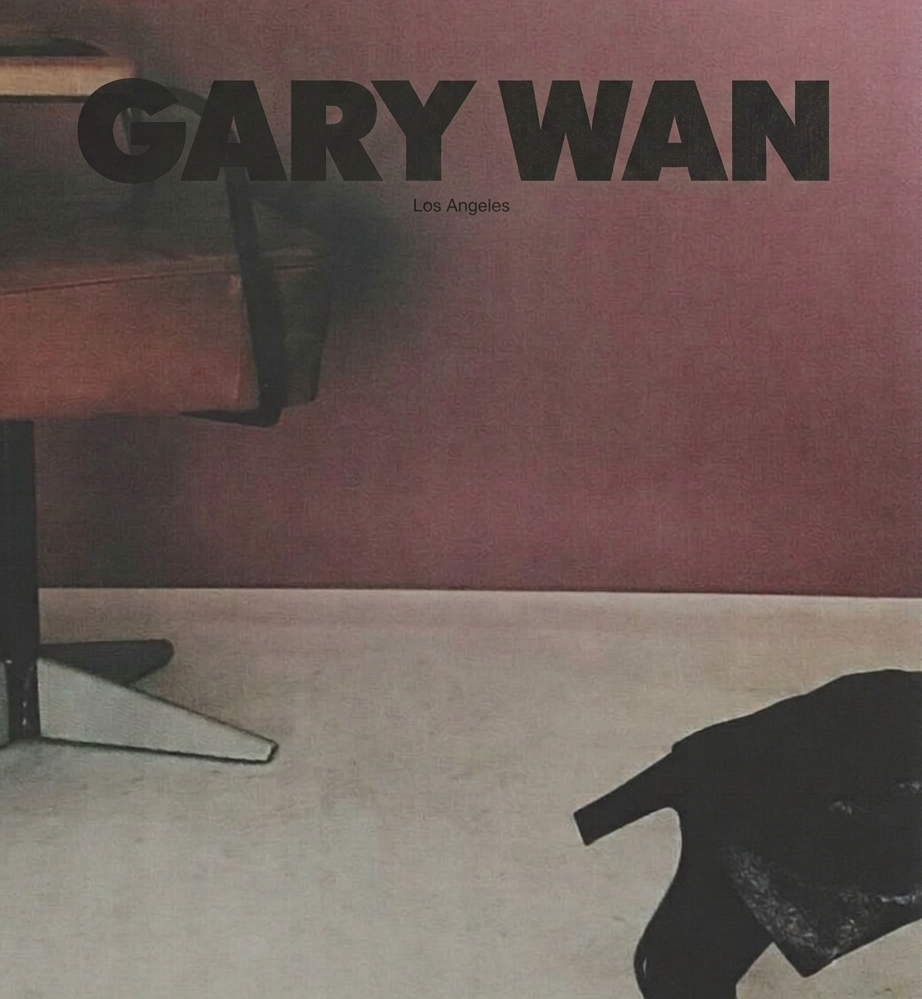 A wall with the text 'GARY WAN Los Angeles' and part of a wooden desk, a black garment or bag on the floor, and part of a black chair or furniture.