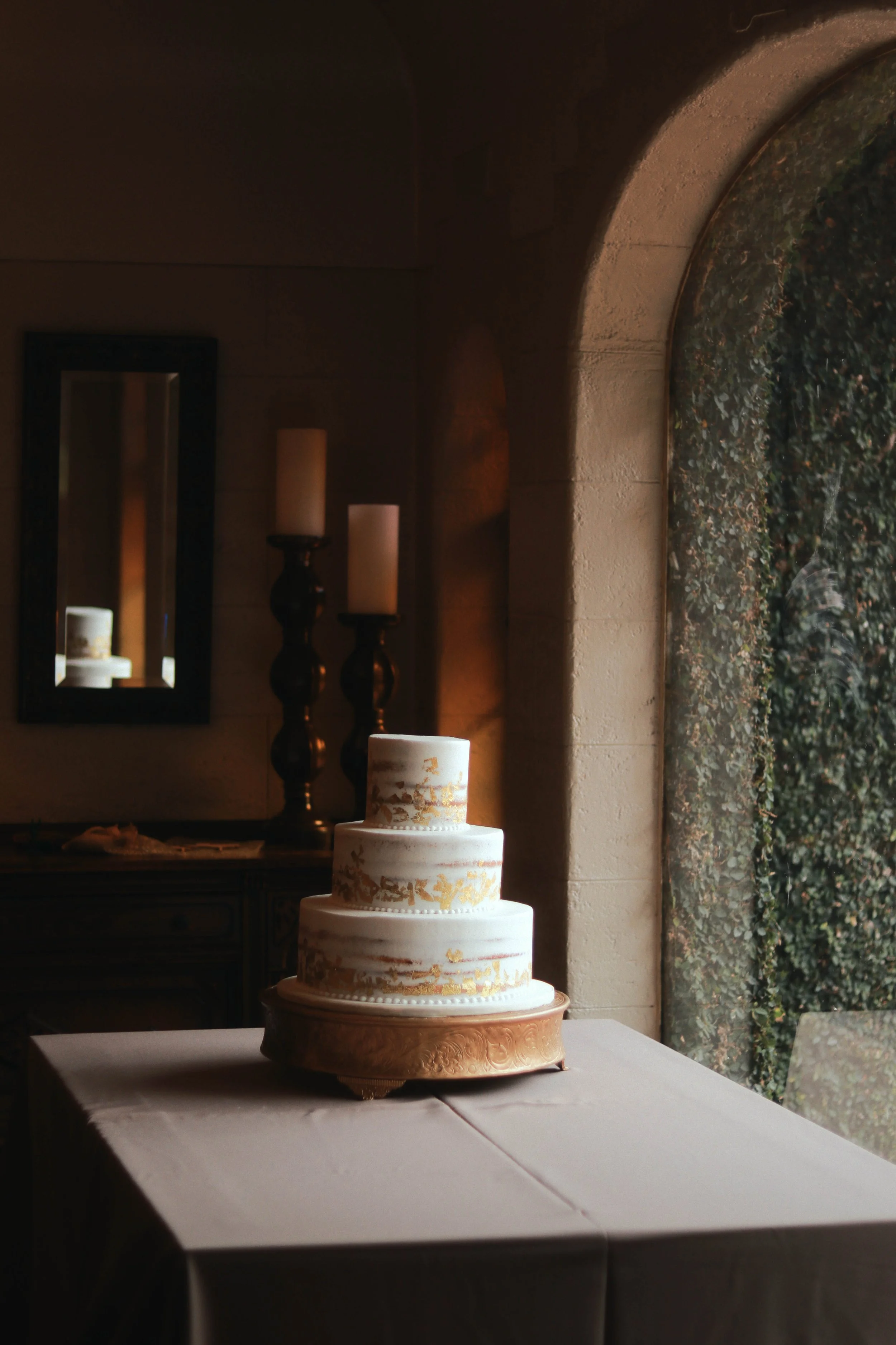 A three-tier white wedding cake with gold decorative accents, placed on a wooden cake stand on a table with a white tablecloth, against a dimly lit background with candles and a mirror.
