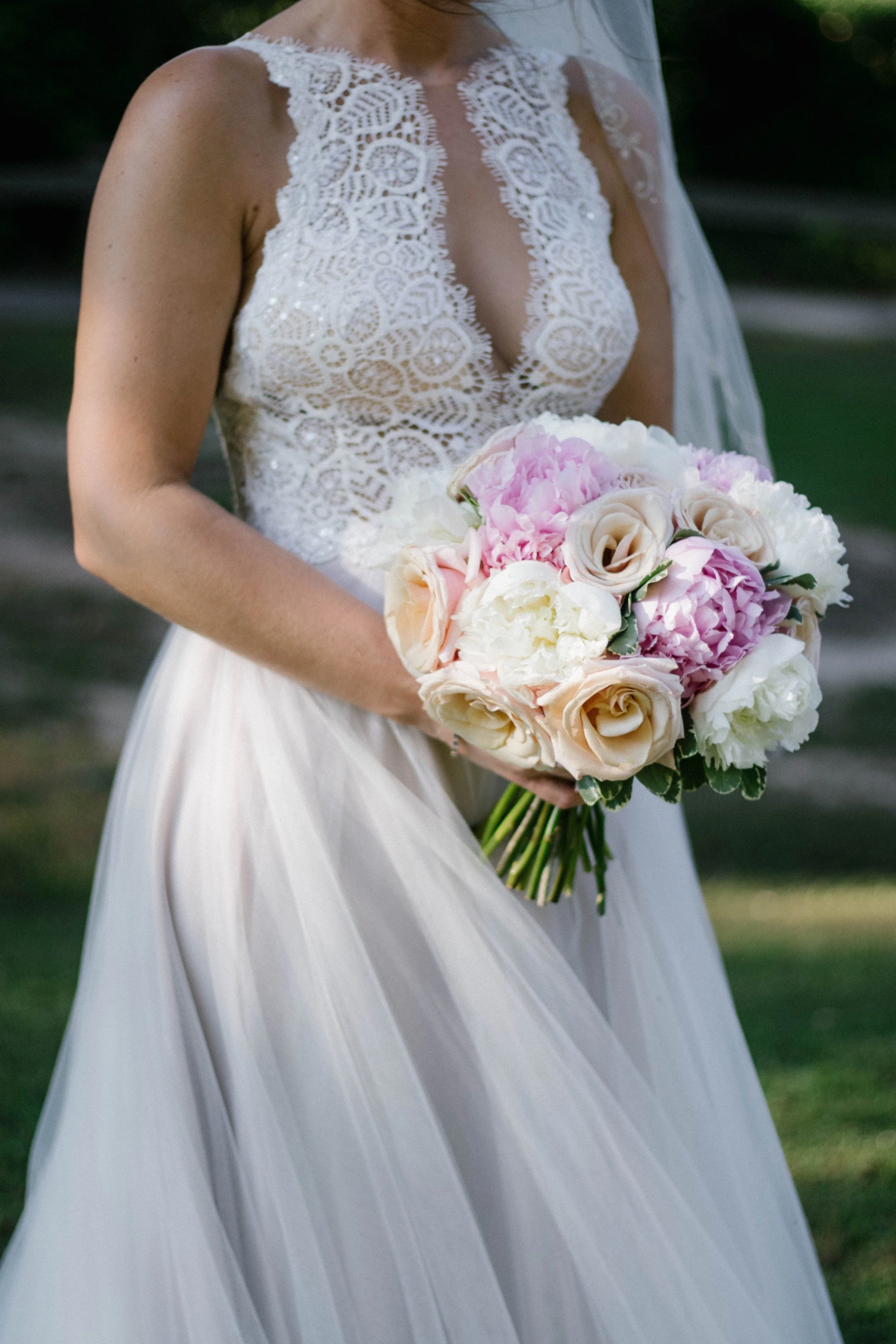 A bride in a lace wedding dress holding a bouquet of pink and white flowers outdoors.