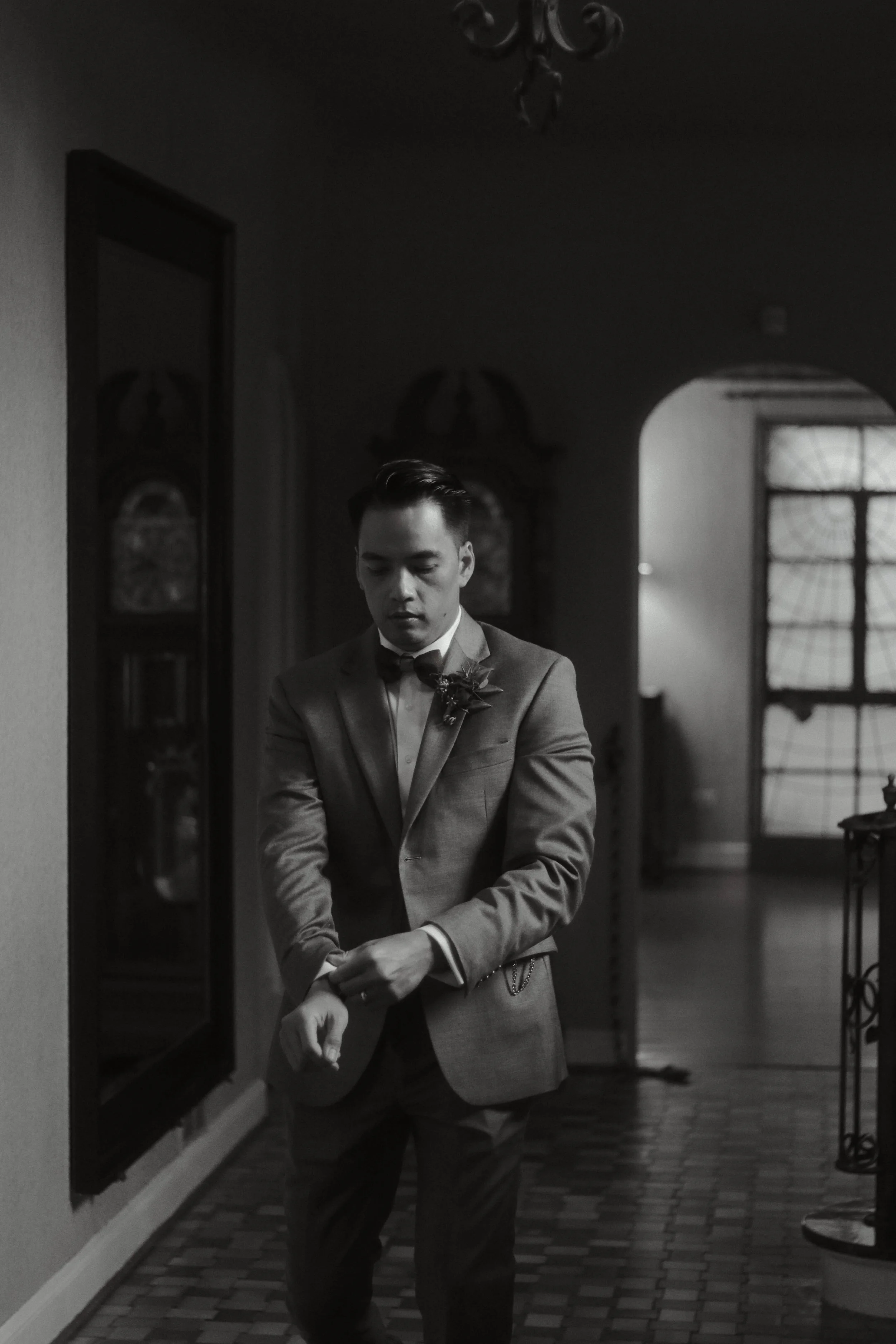 A black and white photo of a man in a suit and bowtie adjusting his cufflinks in an indoor setting with vintage decor.