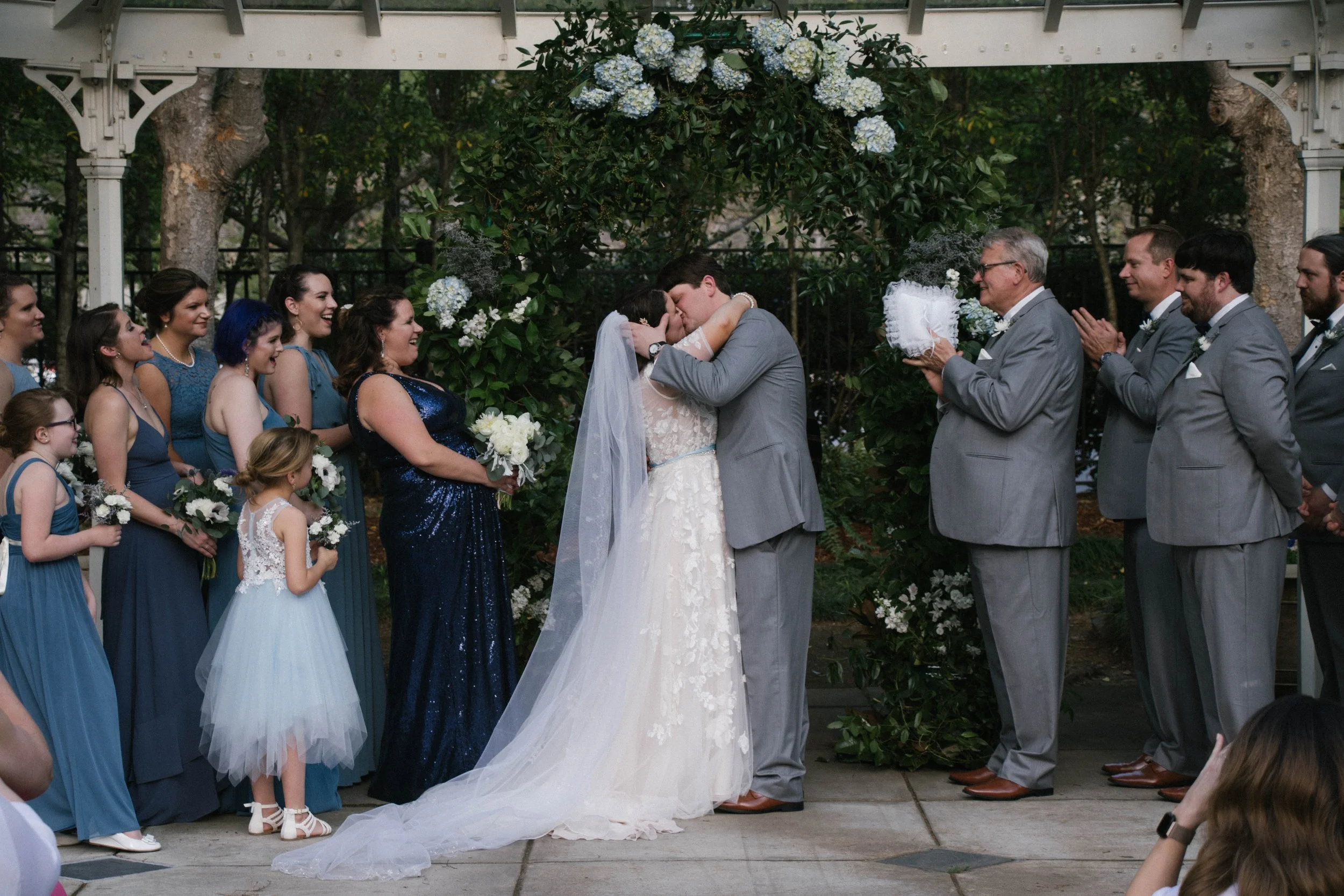 A bride and groom share a kiss during their outdoor wedding ceremony under a floral arch, surrounded by bridesmaids, groomsmen, and children in formal attire.