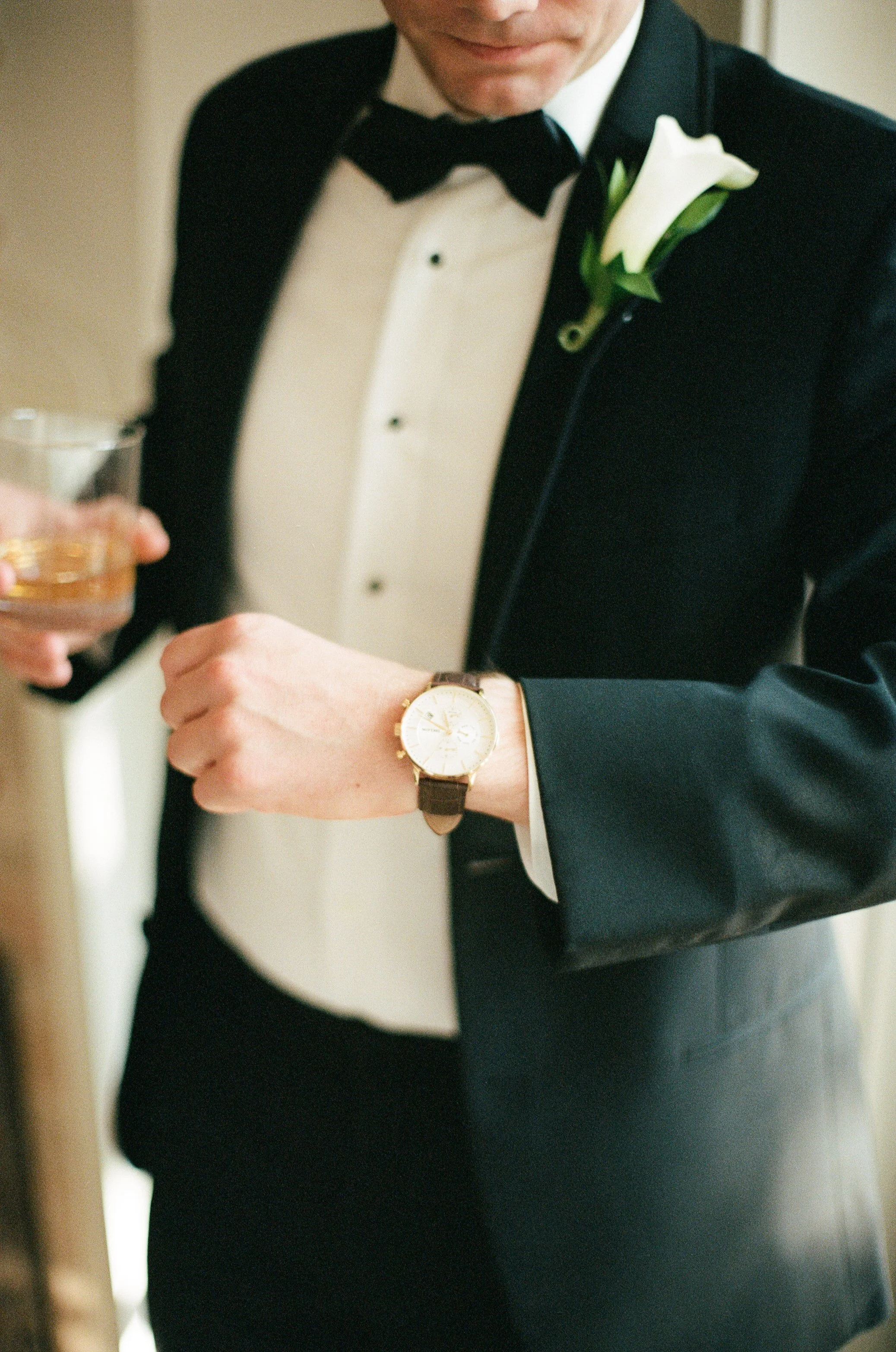 Man in a tuxedo with a boutonniere, checking a watch, holding a drink.