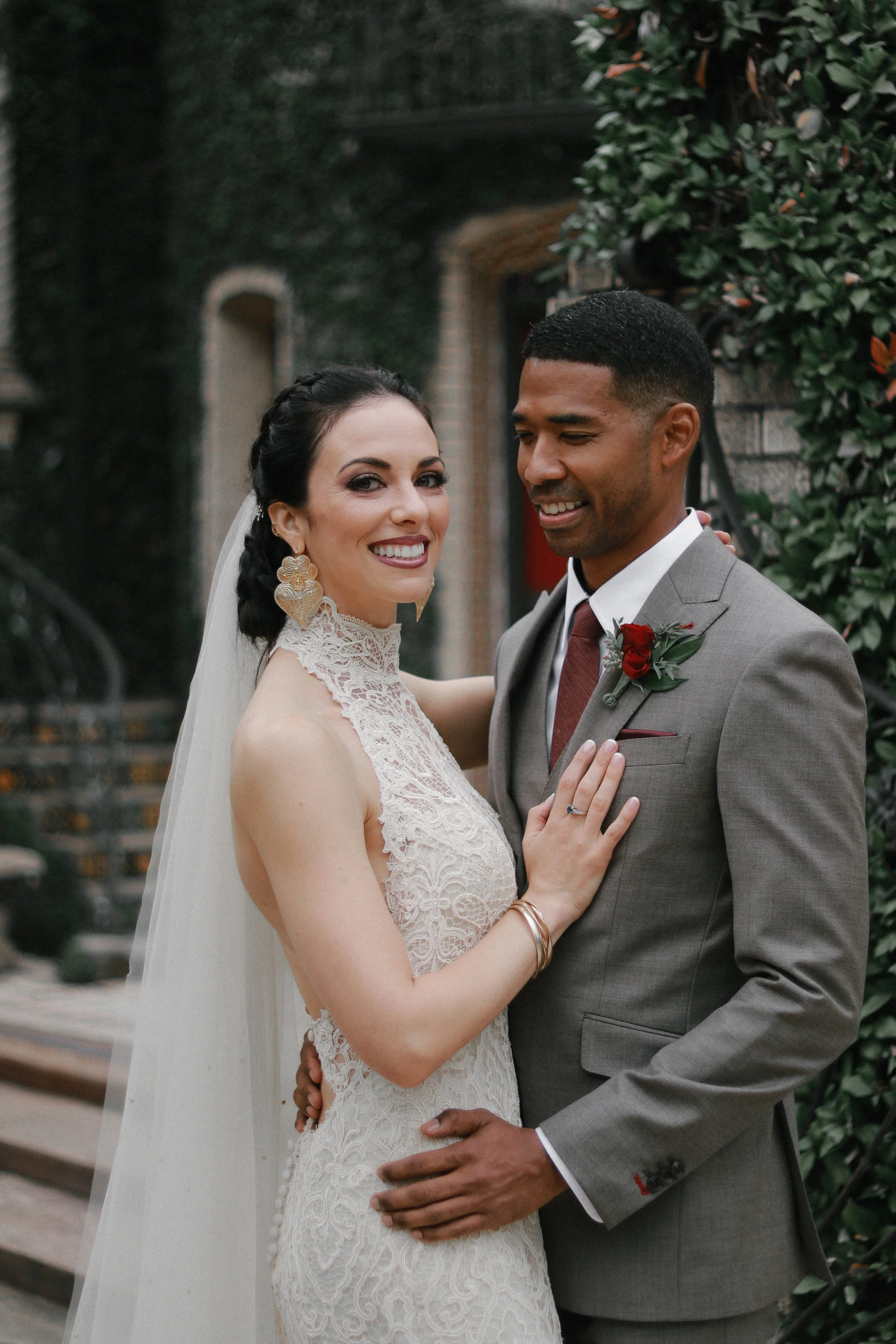 A bride and groom on their wedding day, smiling and embracing outdoors. The bride wears a lace wedding gown with a high neckline and veil, while the groom is dressed in a gray suit with a red boutonniere and a maroon tie. They are standing next to a 