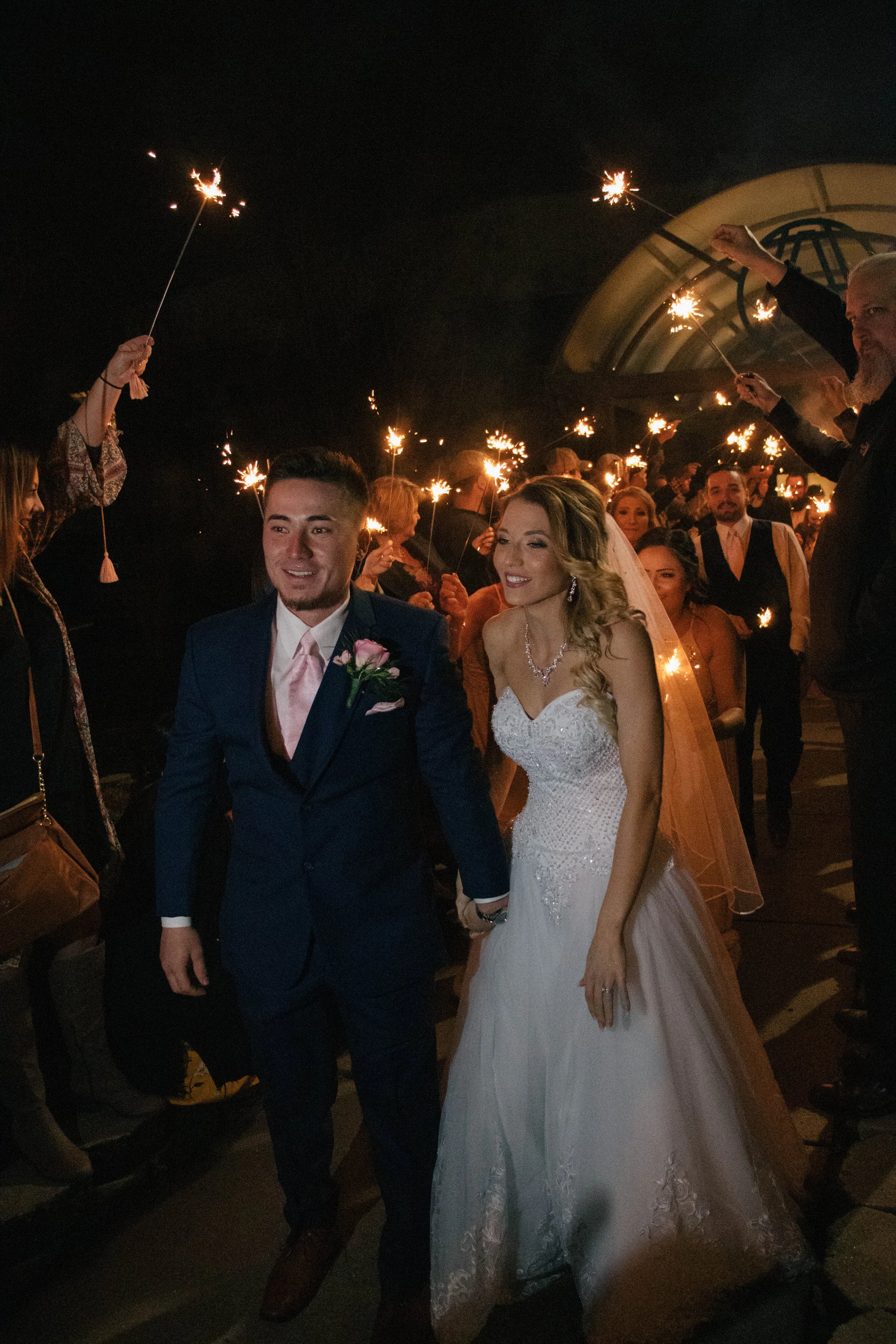 A bride and groom walking outside at night, holding hands, surrounded by guests holding sparklers for a celebration.