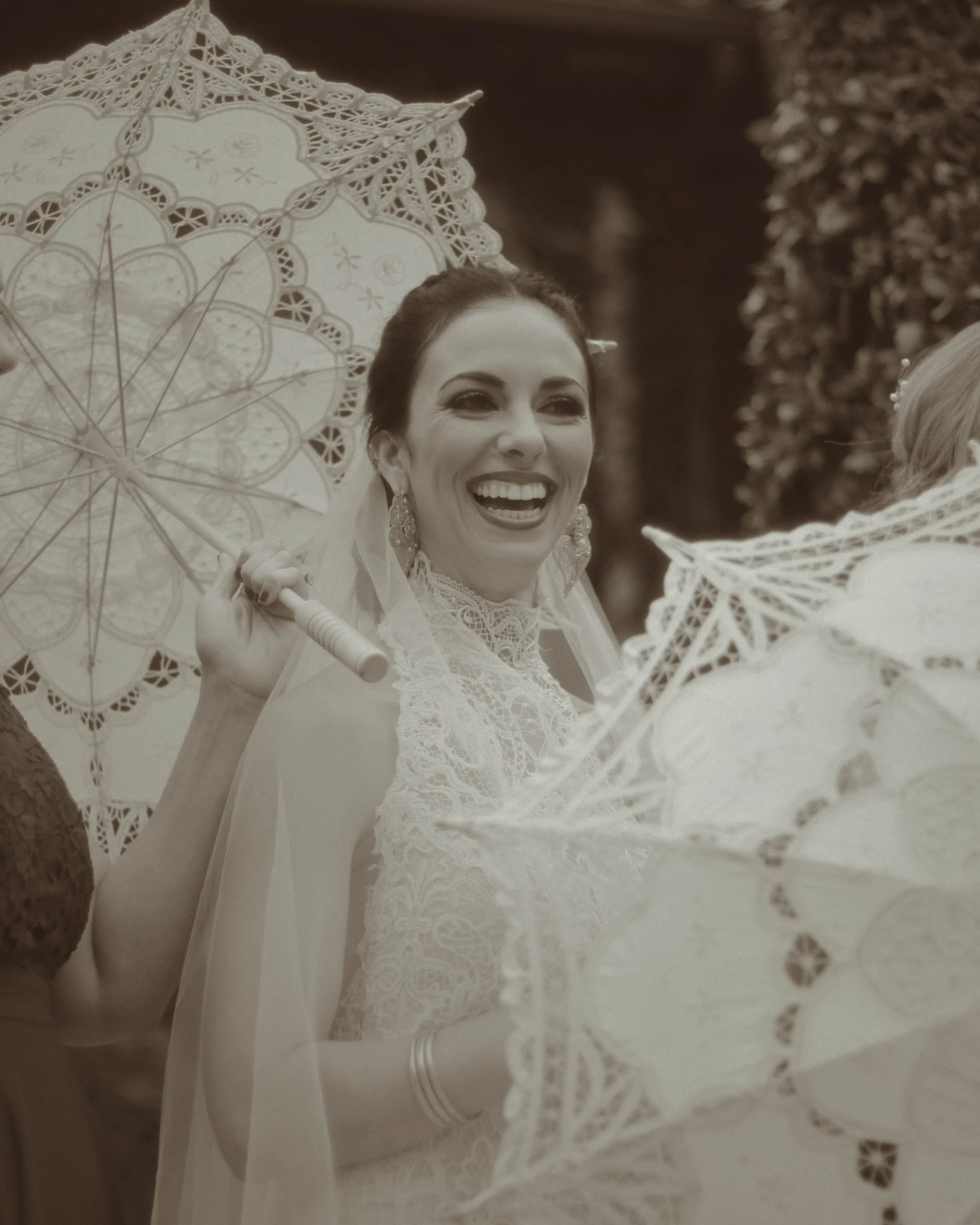 A woman smiling, wearing lace clothing, holding a lace-patterned umbrella, surrounded by similar umbrellas, in an outdoor setting.