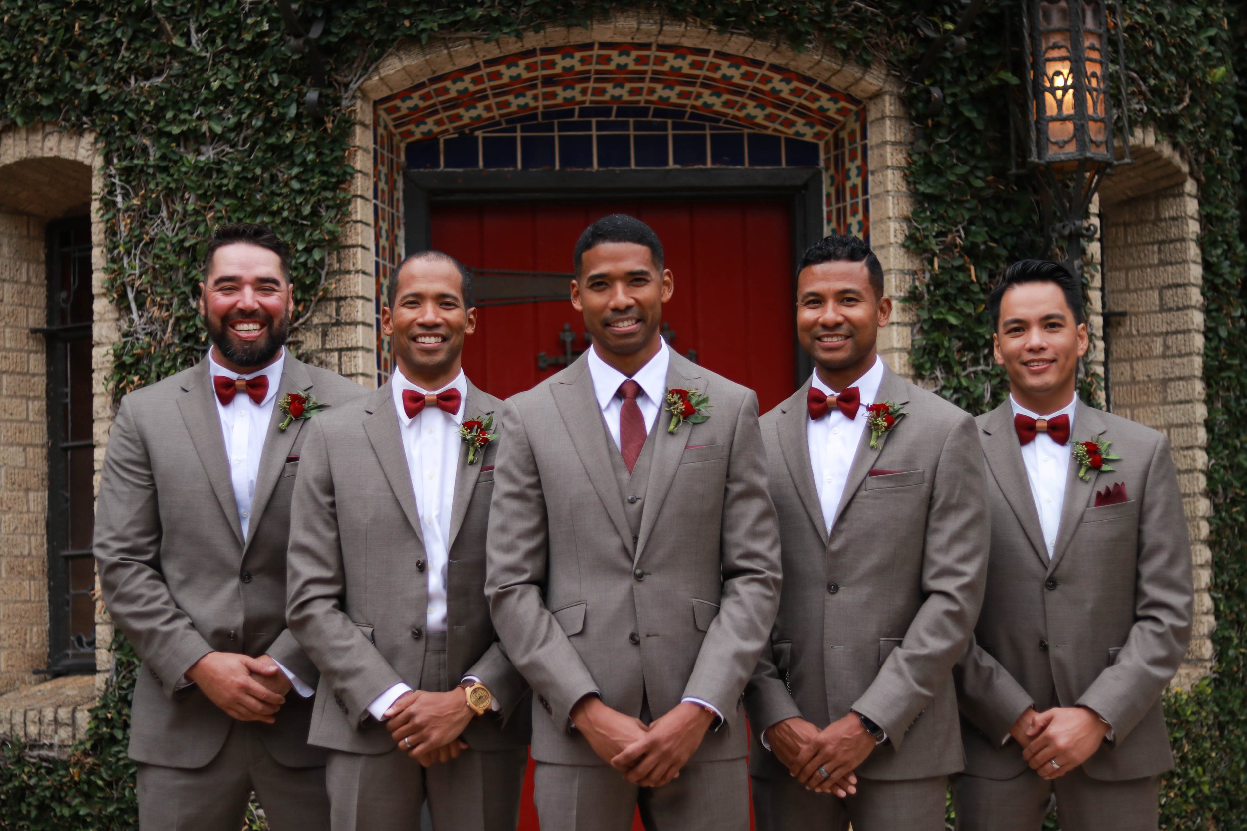 Group of five men in matching beige suits with red bow ties and boutonnières, standing in front of a brick building with a red door, ivy, and ornate tile archway.