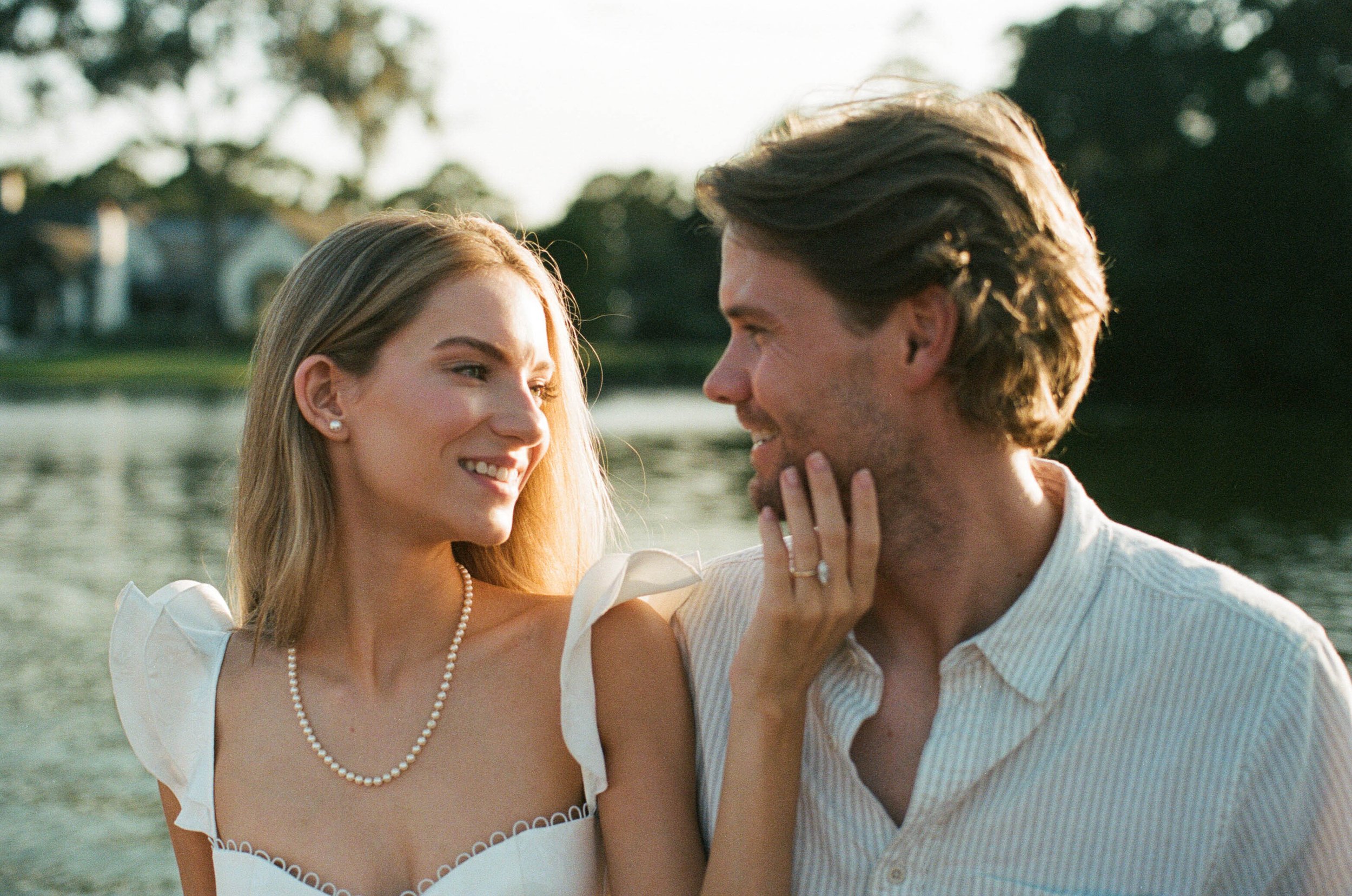 A smiling woman and man facing each other near a body of water, with trees and houses in the background, during sunset.
