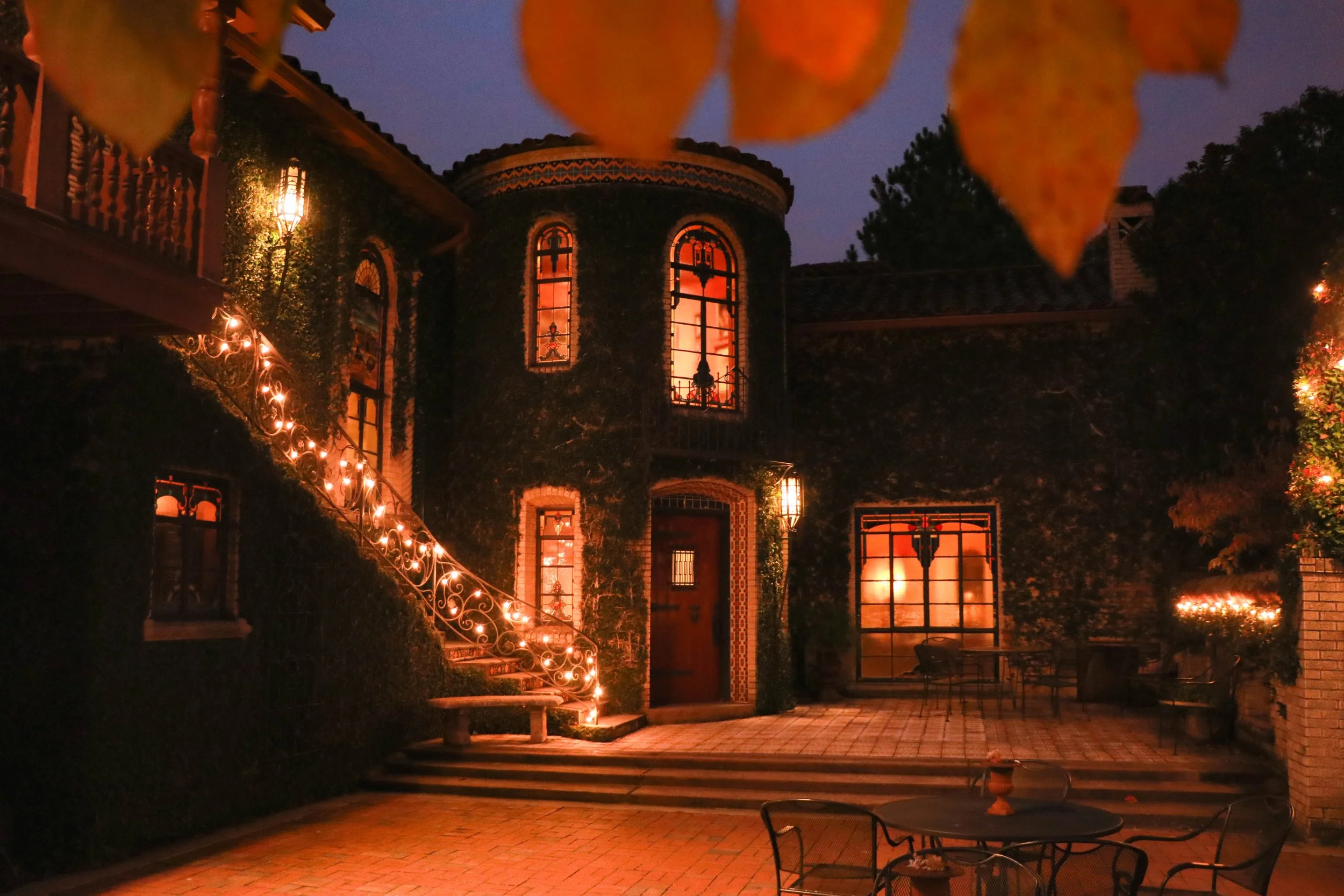 Nighttime view of a house with ivy-covered walls, illuminated by warm lights, with a staircase decorated with string lights, and an outdoor patio with chairs and tables.