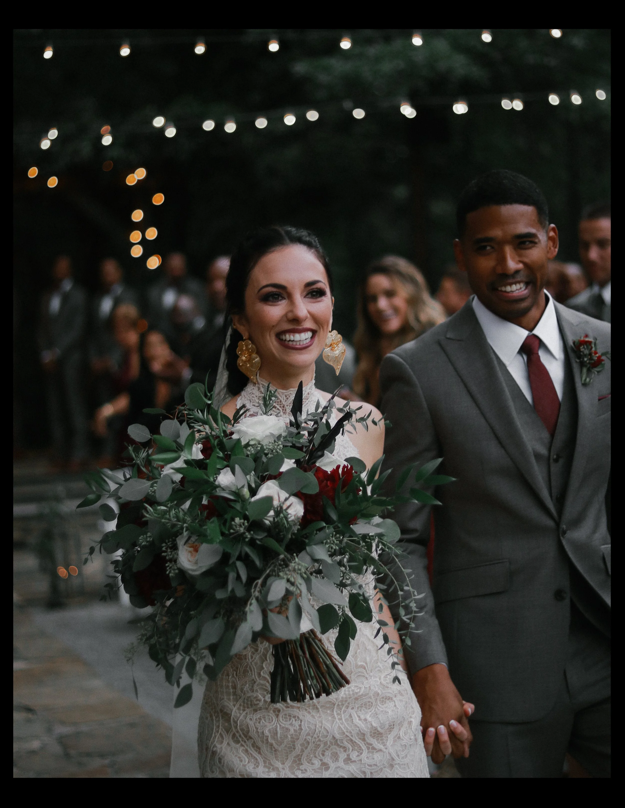 A newlywed couple walking down the aisle holding hands, surrounded by guests and string lights, celebrating their wedding outdoors at night.