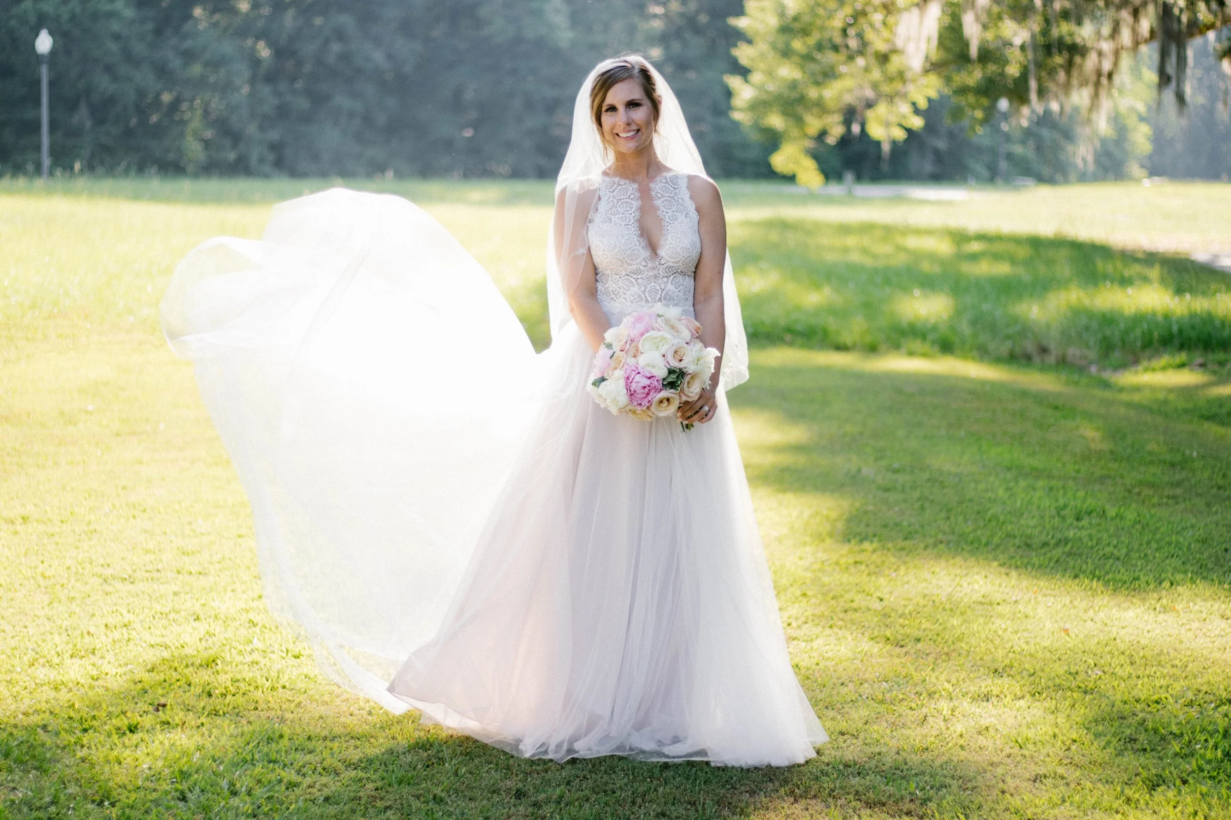 A bride in a white wedding gown holding a bouquet of pink and white flowers standing on a grassy field with trees in the background.