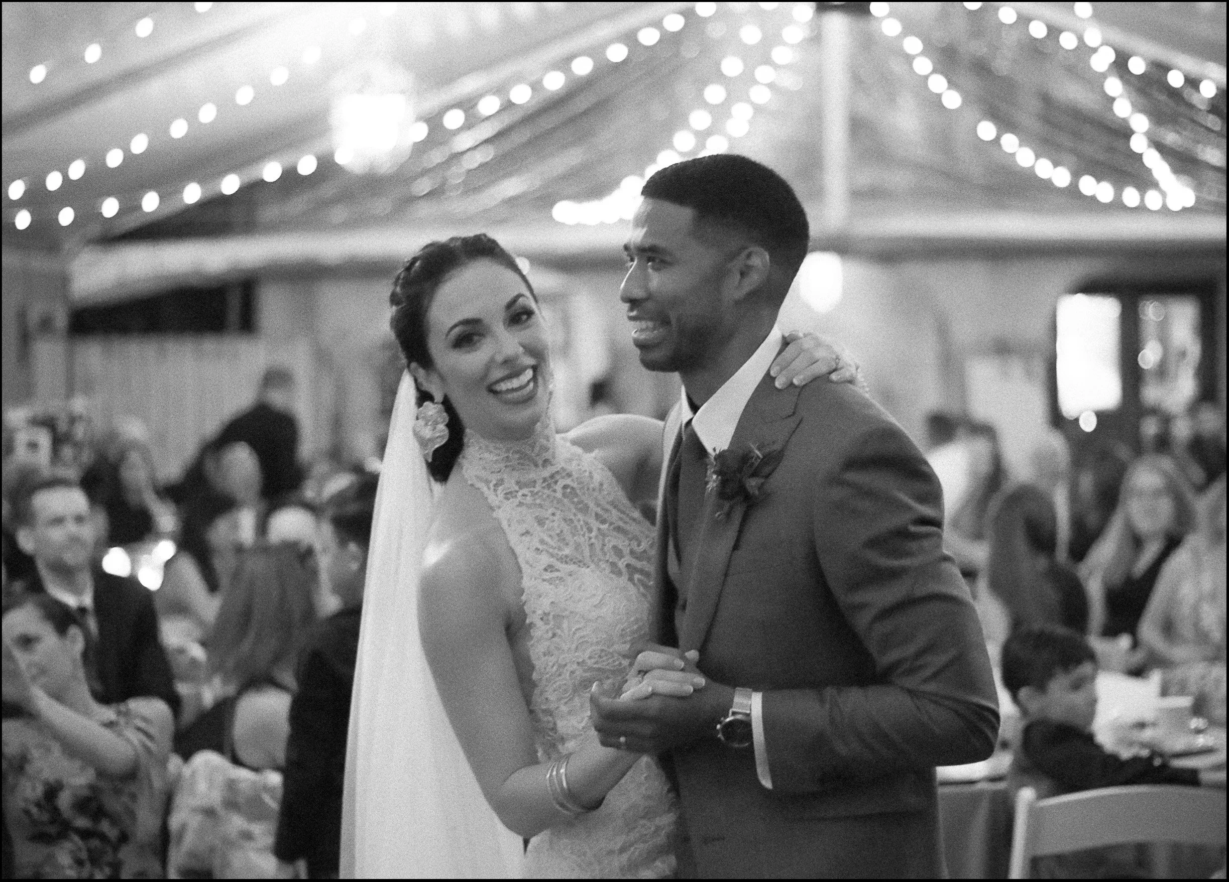 Black and white photo of a bride and groom dancing at their wedding reception, surrounded by guests, under decorative string lights.