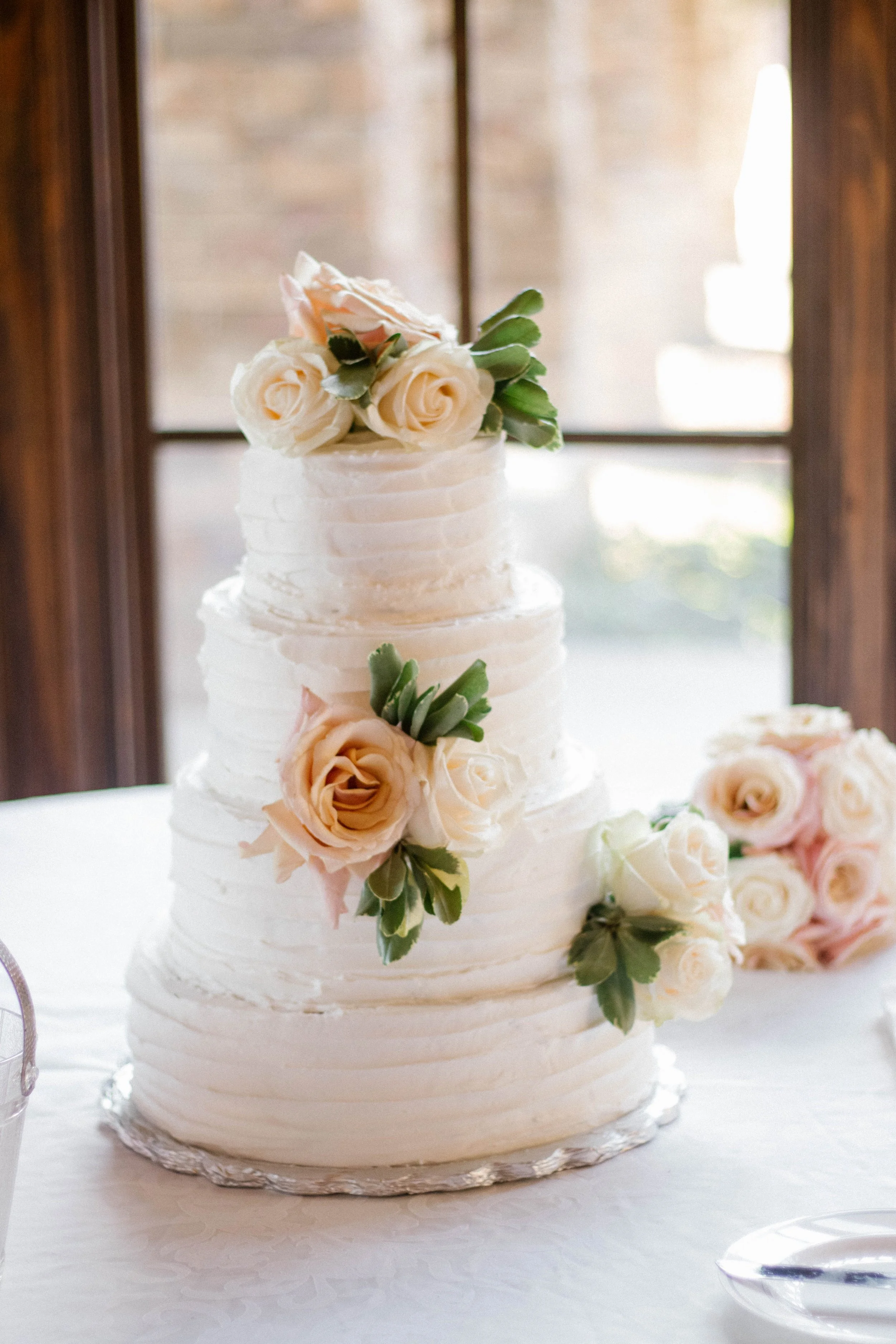 Three-tier white wedding cake decorated with cream-colored roses and green leaves, sitting on a silver cake board.