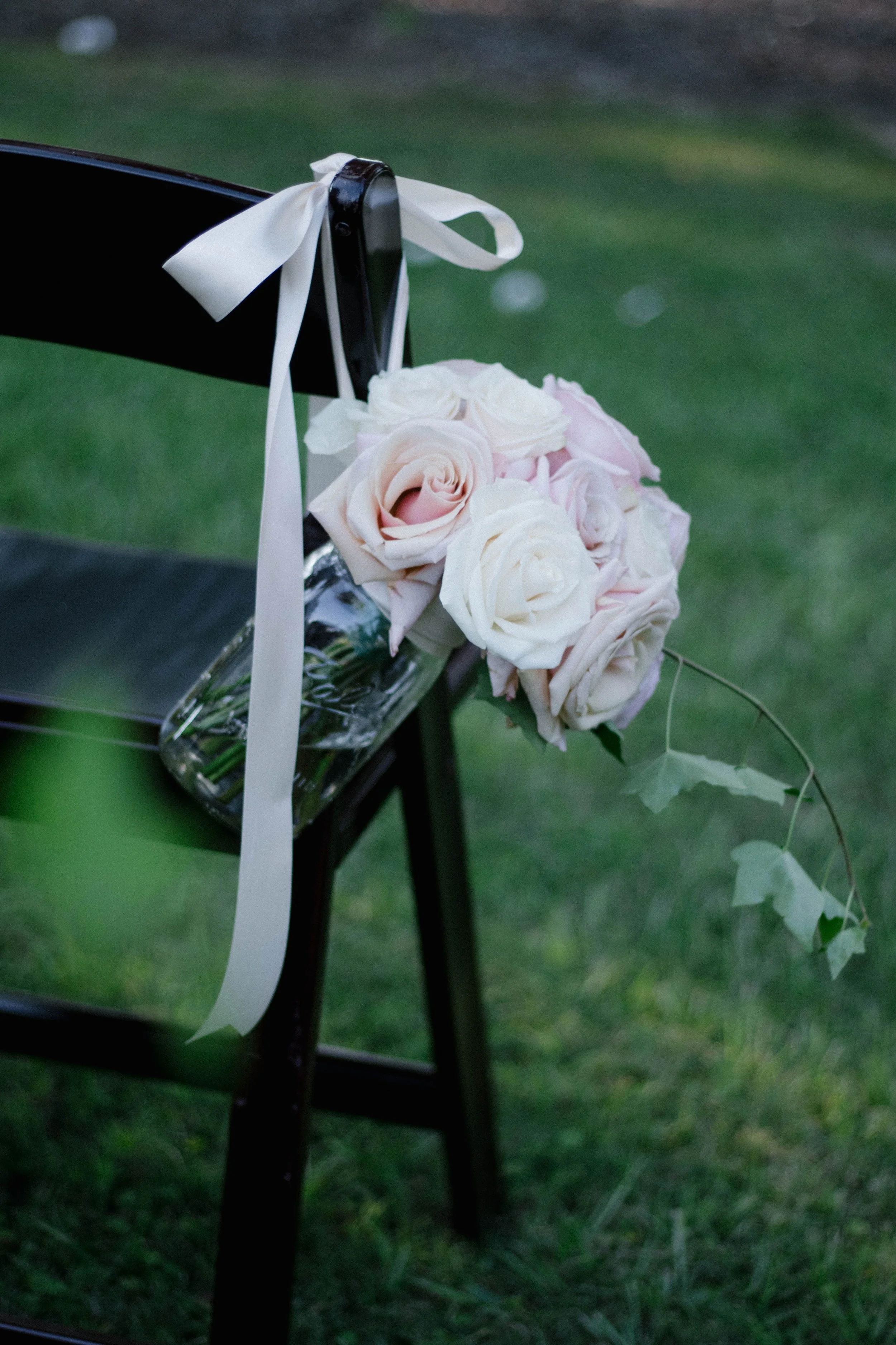 A bouquet of pink and white roses with green leaves attached, resting on a black chair with a white ribbon tied to the chair's backrest, set on grass.