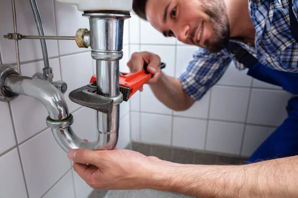 Licensed plumber repairing a leaking drain pipe beneath a bathroom sink, highlighting the importance of addressing water issues before cosmetic home renovations.