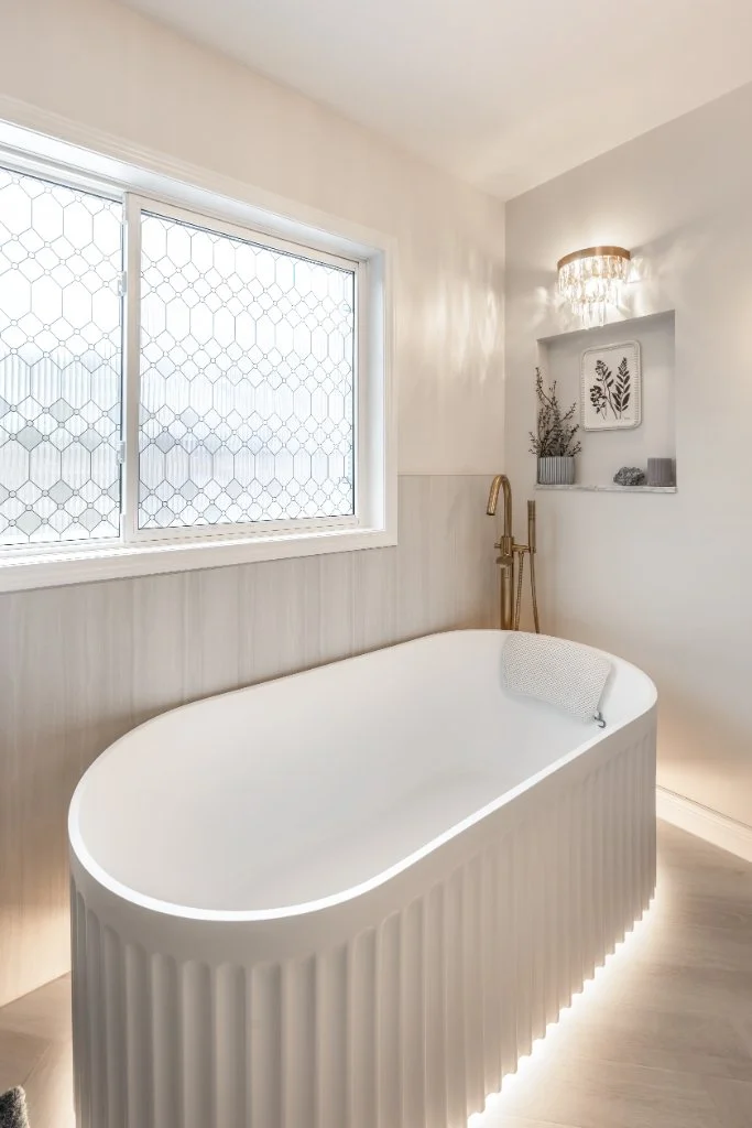 Modern oval freestanding tub with integrated LED lighting, gold floor-mounted faucet, and custom wall niche in a luxury ensuite bathroom.