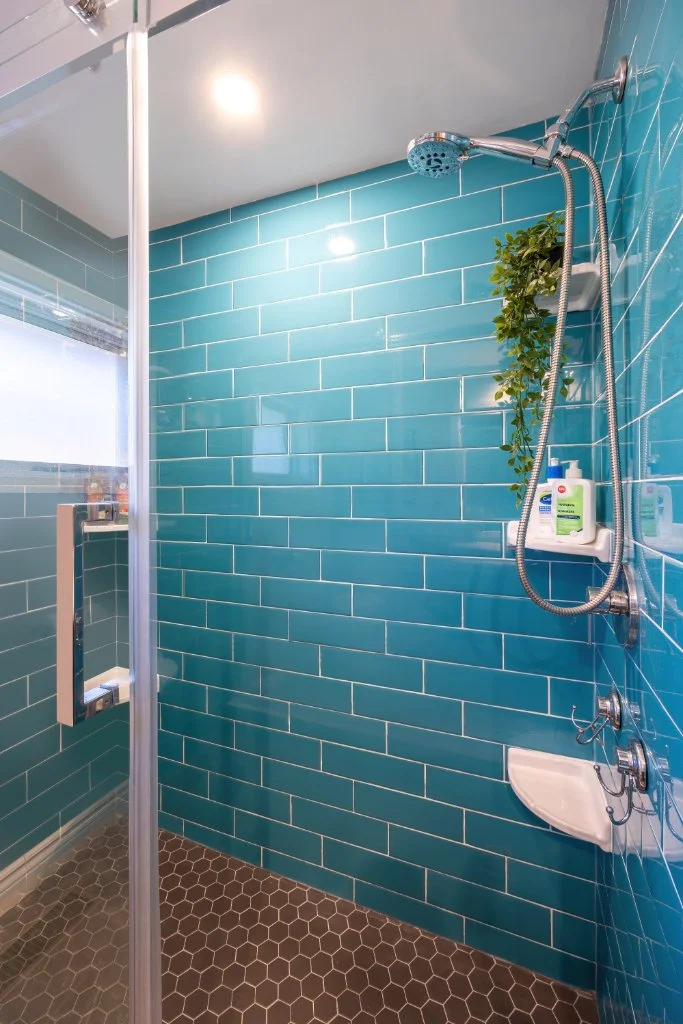 Walk-in shower with bright blue subway tile, handheld showerhead, corner shelf and dark hexagon floor tile.