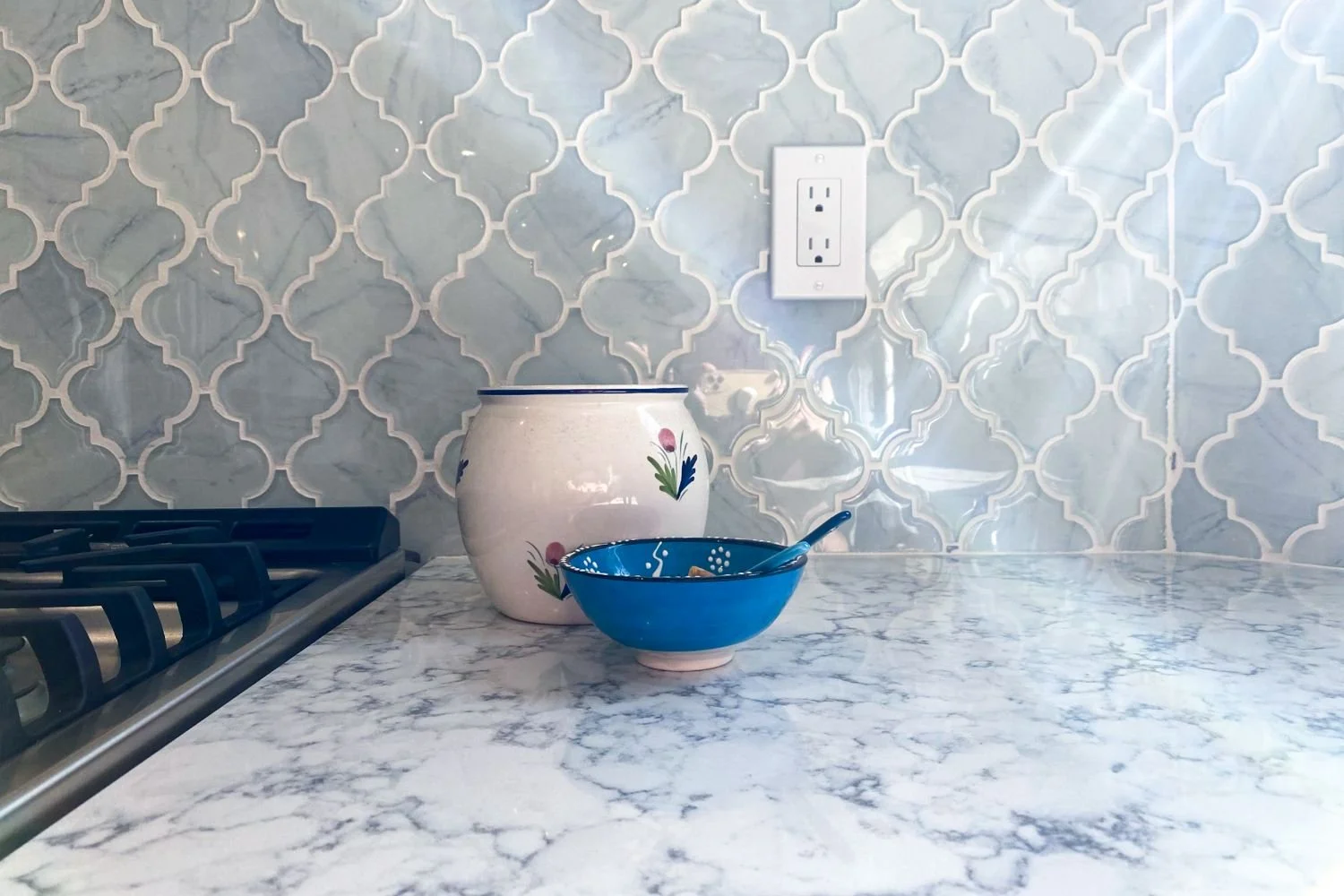 A close-up view of a light blue arabesque tile backsplash with a marble-look countertop, featuring a ceramic jar and blue bowl beside a gas cooktop.