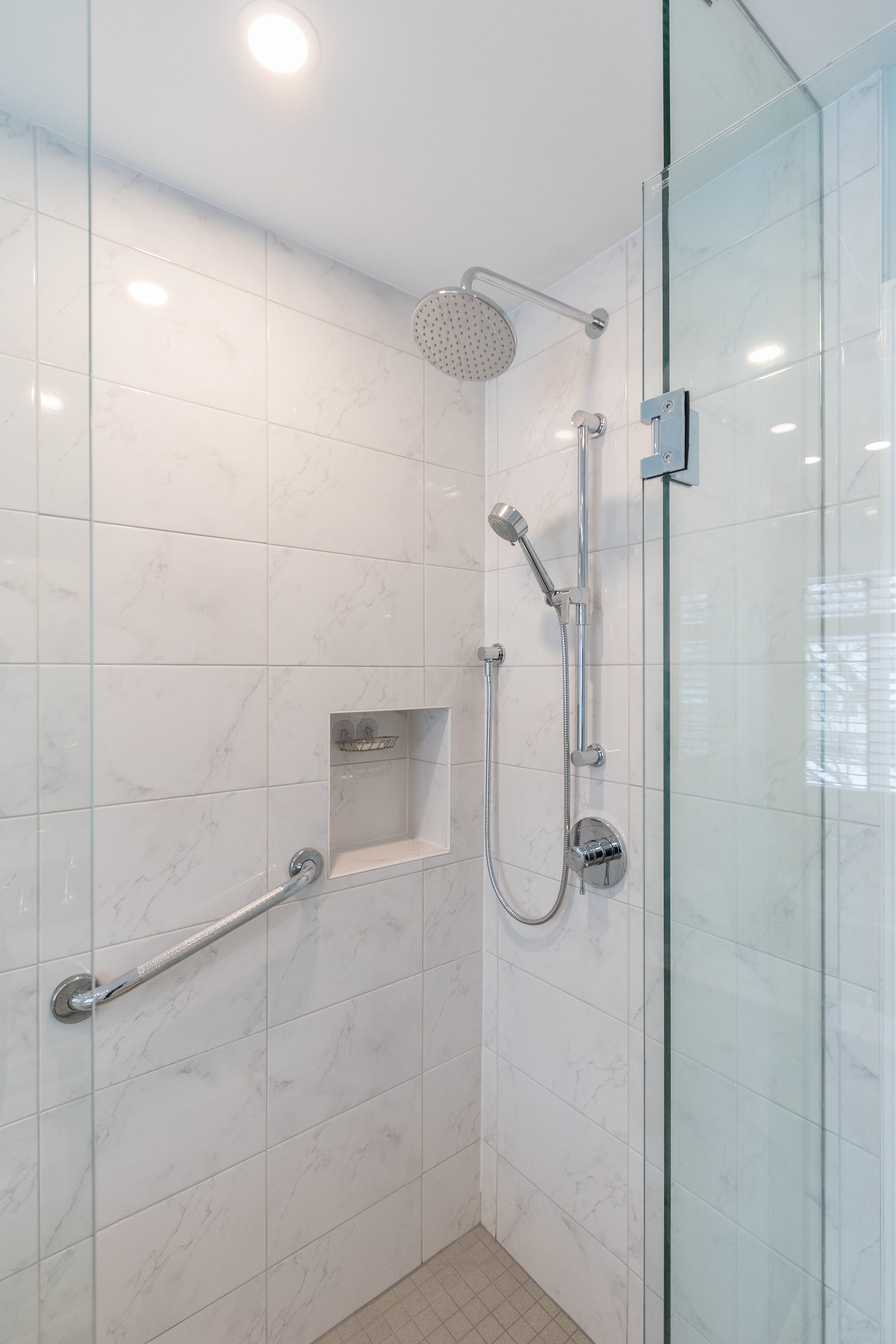 Marble-look porcelain tile shower with frameless glass, built-in niche and grab bar combining style with accessibility.
