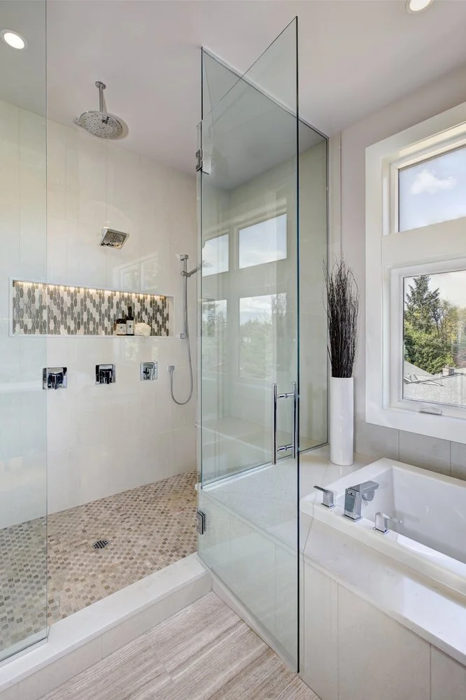 Modern bathroom renovation featuring a walk-in glass shower with custom tile niche, rainfall shower head, and freestanding bathtub beside large windows.