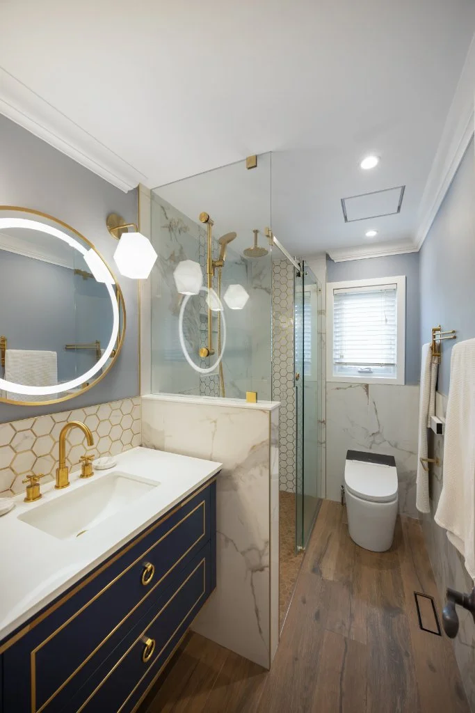 Gorgeous renovated bathroom in North Whitby with blue and gold tones. Large 12x24 tiles line the shower walls with an accent tile with white hexagon tiles outlined in gold. A lighted mirror at the wall hung navy blue vanity with a gold faucet adds.