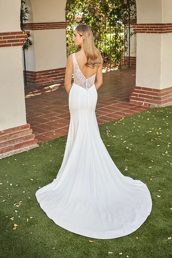 Back view of a crepe fit-and-flare wedding dress featuring a deep illusion beaded V-back, fabric-covered buttons, and a graceful chapel train, photographed in a romantic outdoor courtyard.