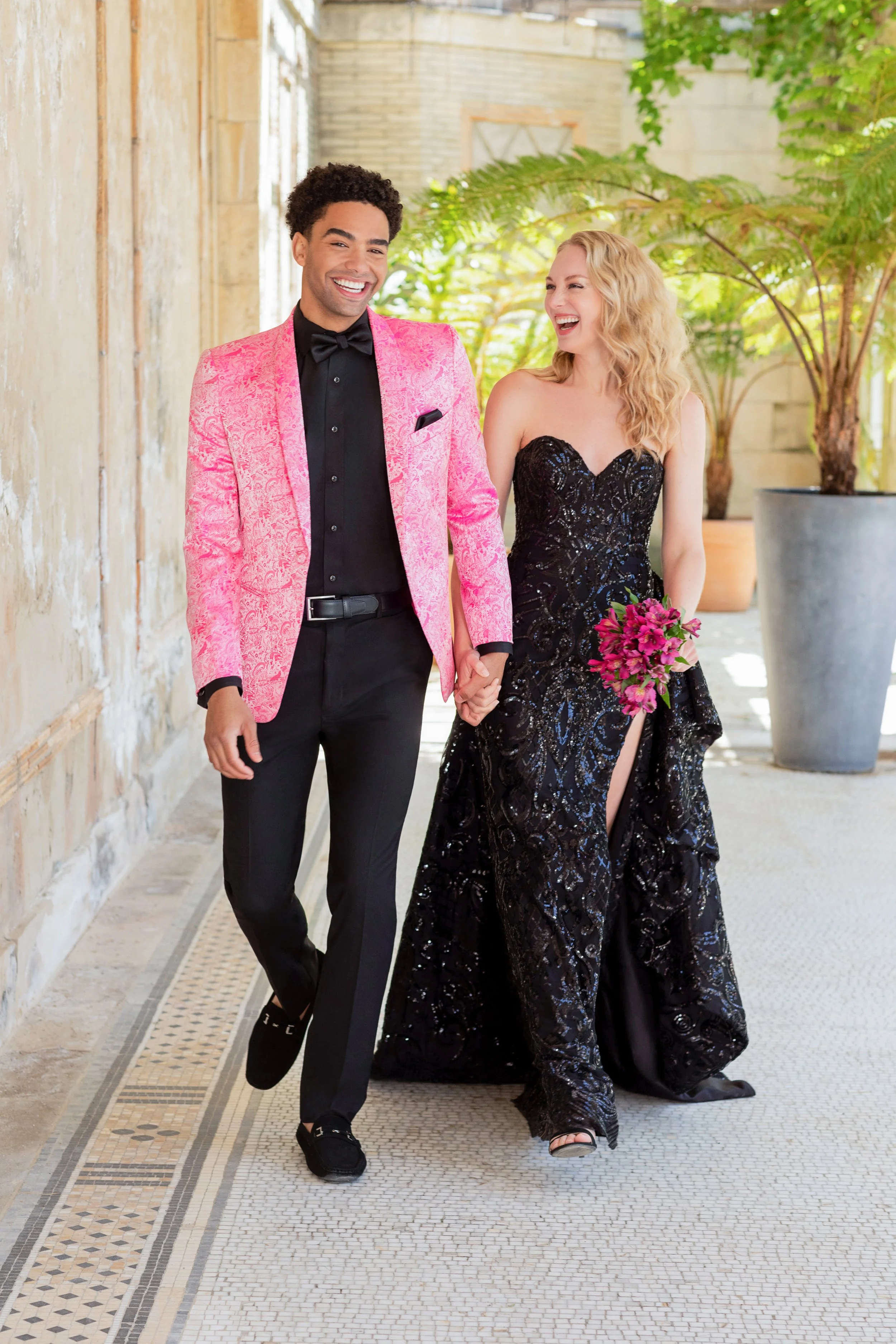 Prom couple strolls hand in hand in coordinated formalwear, featuring a bold pink patterned tuxedo jacket and a sparkling black sequined gown with a sweetheart neckline and high slit, creating a stylish and modern prom portrait.
