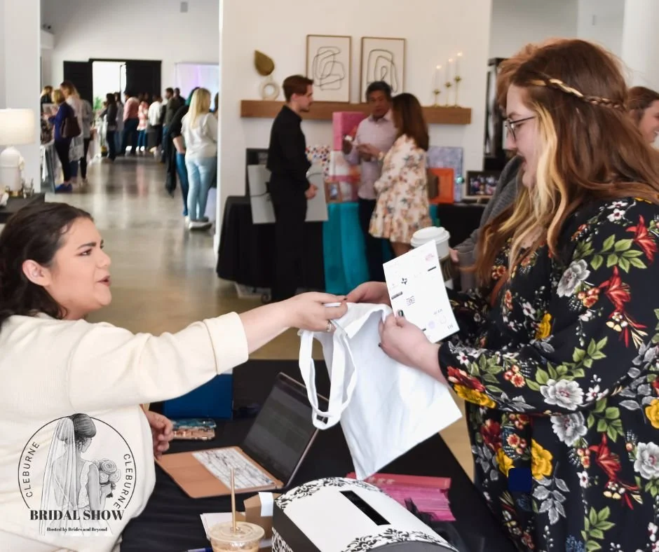 Attendee receives a welcome tote bag and event materials at a bridal show registration table, with wedding vendors and guests gathering throughout the event space in the background.