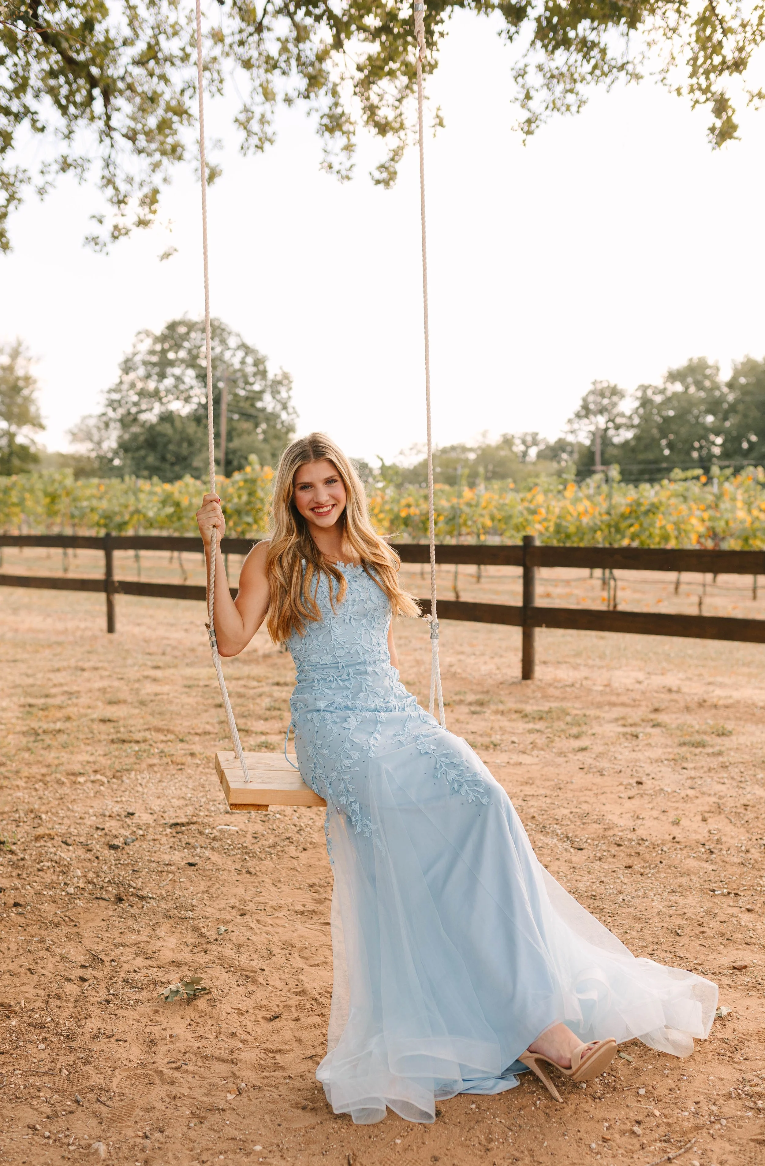 A smiling young woman sits on a wooden swing wearing a flowing light blue formal gown with delicate floral details. The outdoor setting, framed by trees and a rustic fence, creates a charming prom or special occasion portrait.
