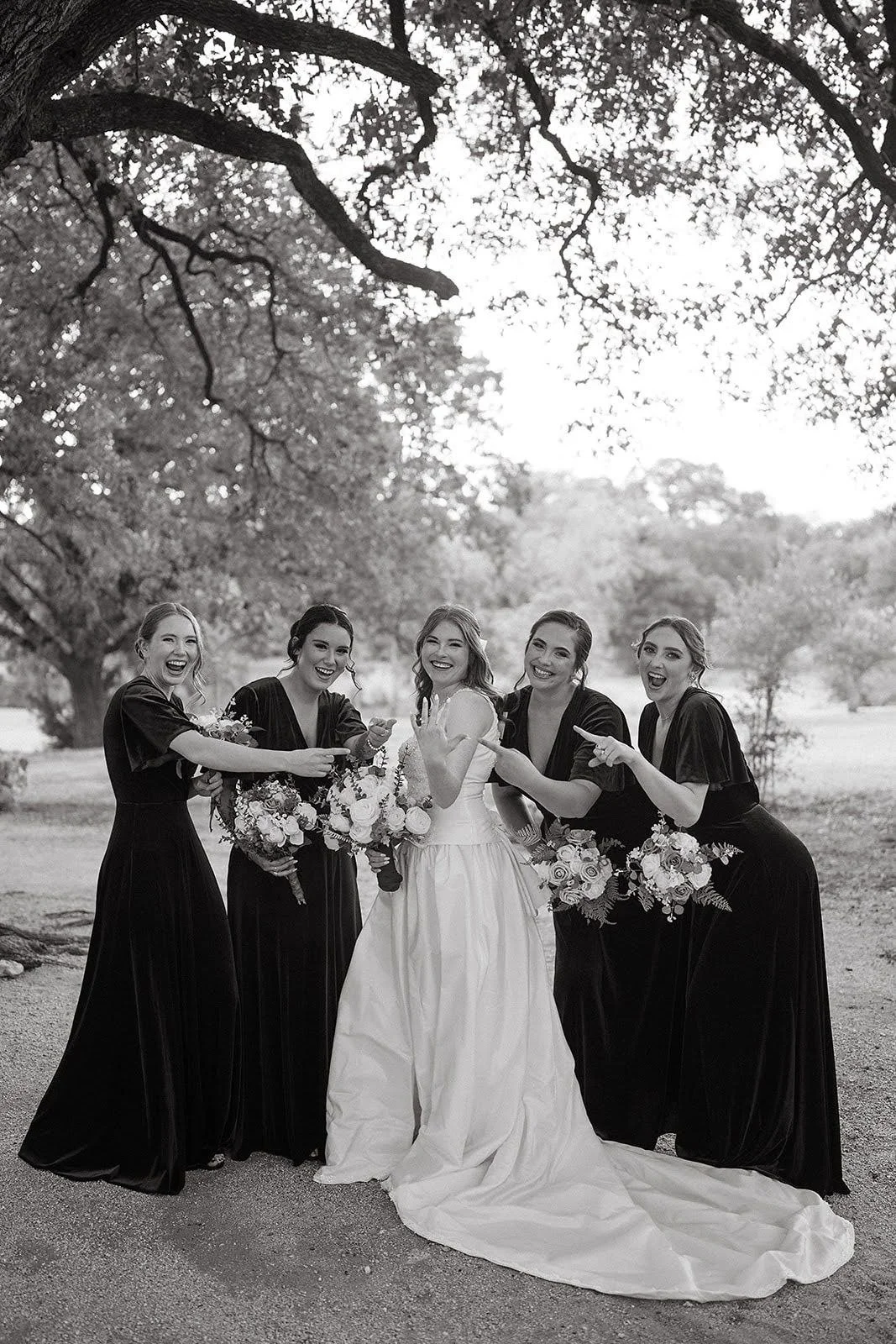 Bride in a classic white gown shares a playful moment with four bridesmaids in coordinating dark dresses, holding floral bouquets and laughing together during a joyful outdoor wedding portrait.