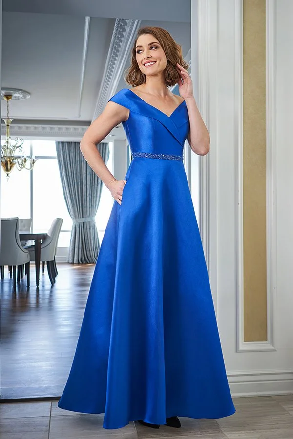 Woman modeling a royal blue off-the-shoulder mother of the bride gown featuring an A-line silhouette and beaded waistband in an elegant indoor setting.