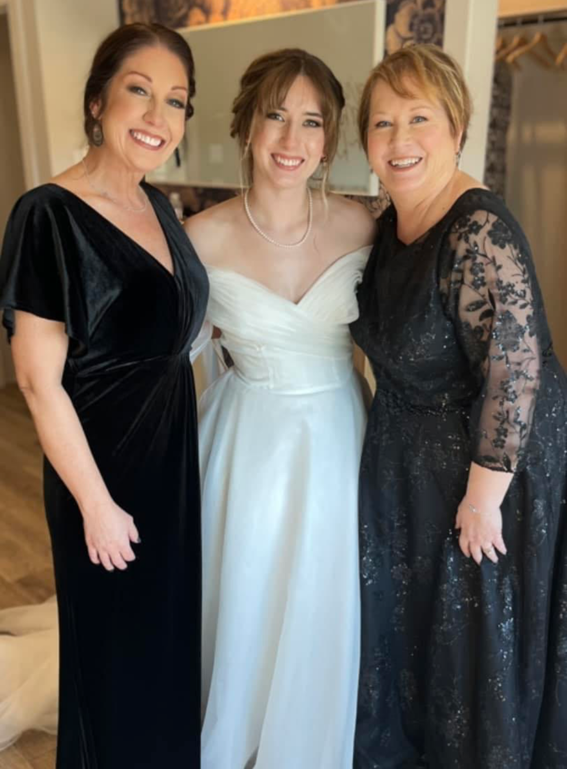A bride stands smiling between two women in elegant black gowns, all posing together in a bright dressing room before the wedding ceremony.
